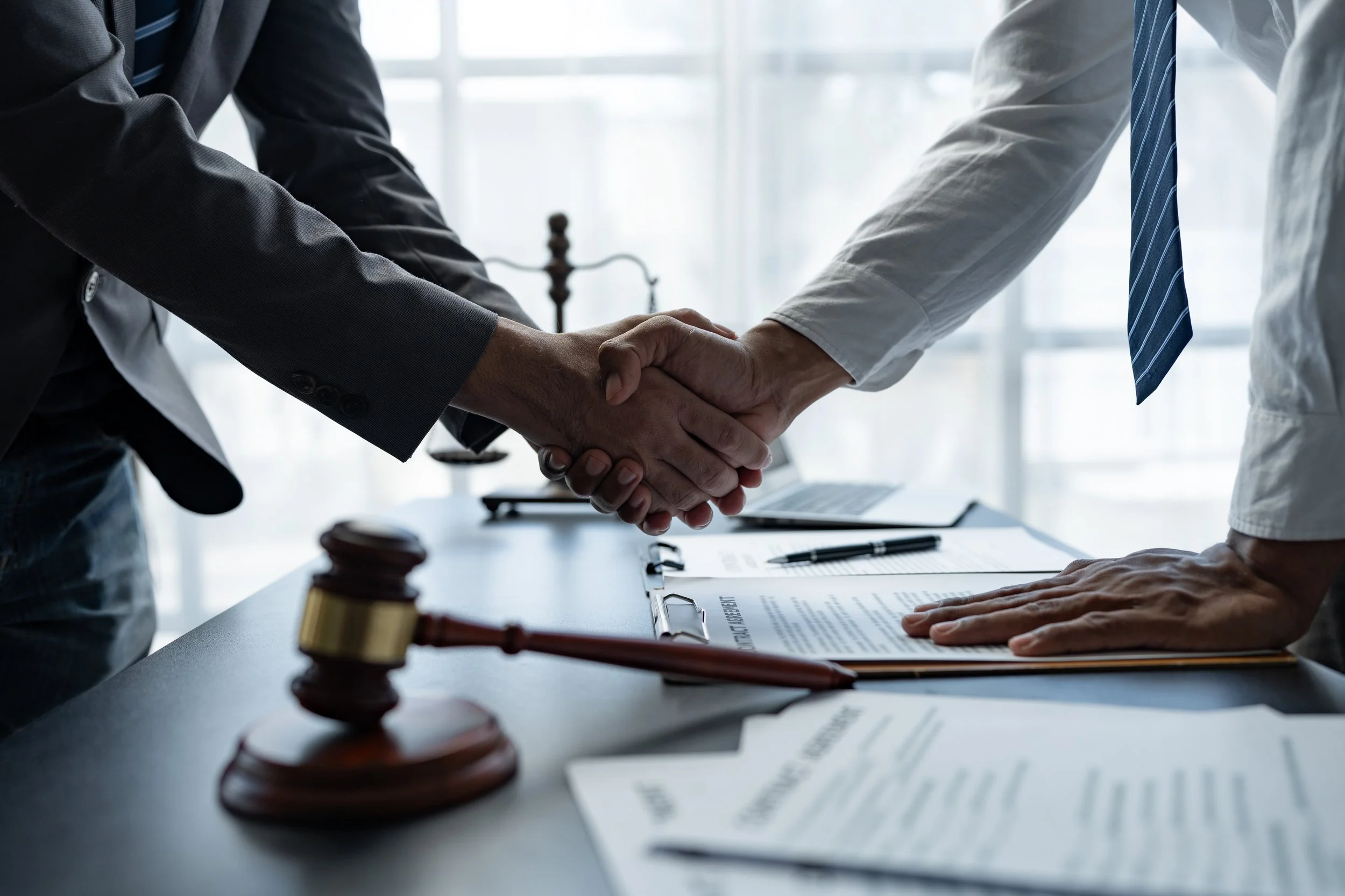 Two individuals in business attire shaking hands across a table with legal documents and gavel, indicating a formal agreement or contract signing.