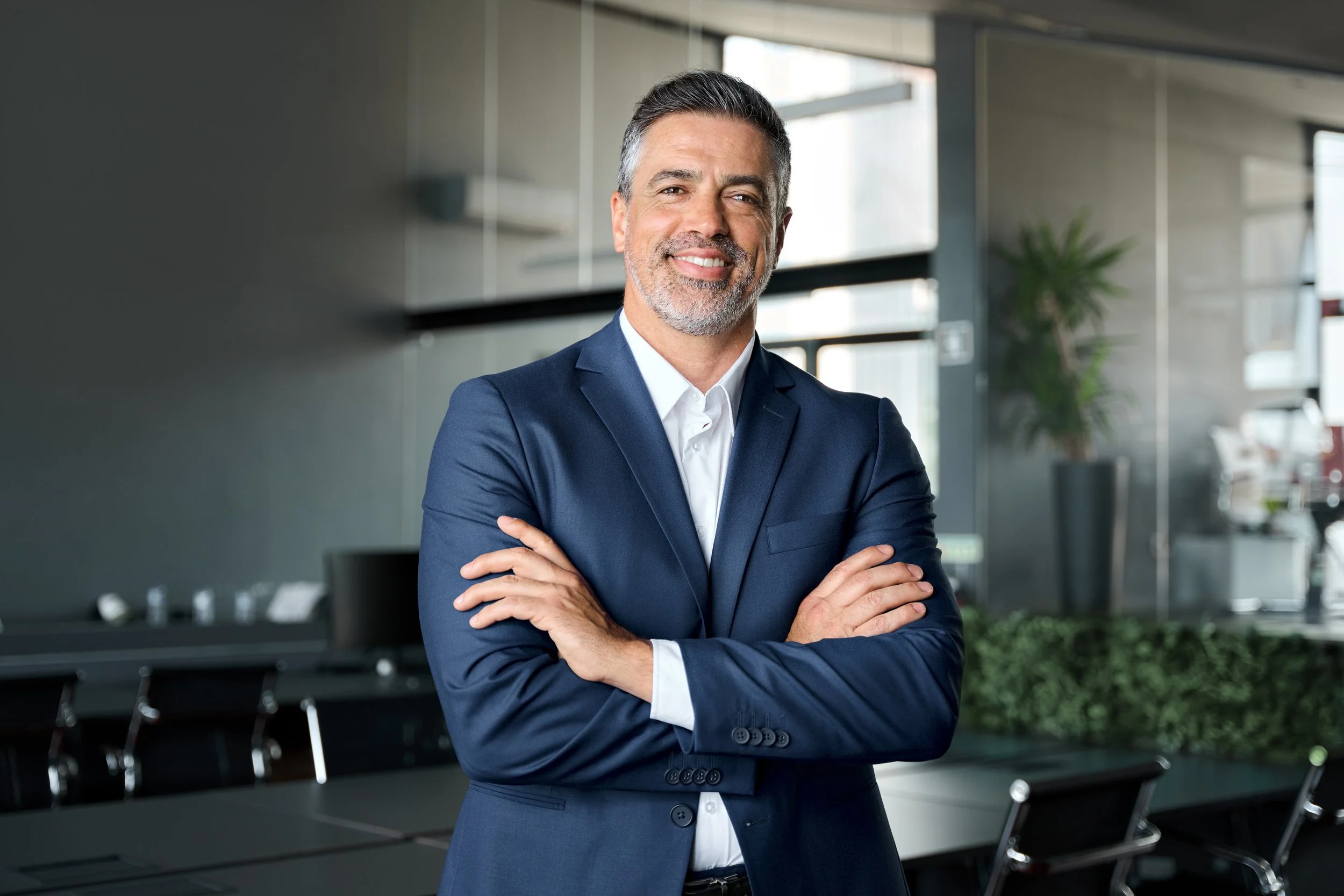 A middle-aged man with gray hair and a beard, dressed in a navy blue suit with a white shirt, standing with arms crossed in a modern office space with large windows, plants, and conference tables.