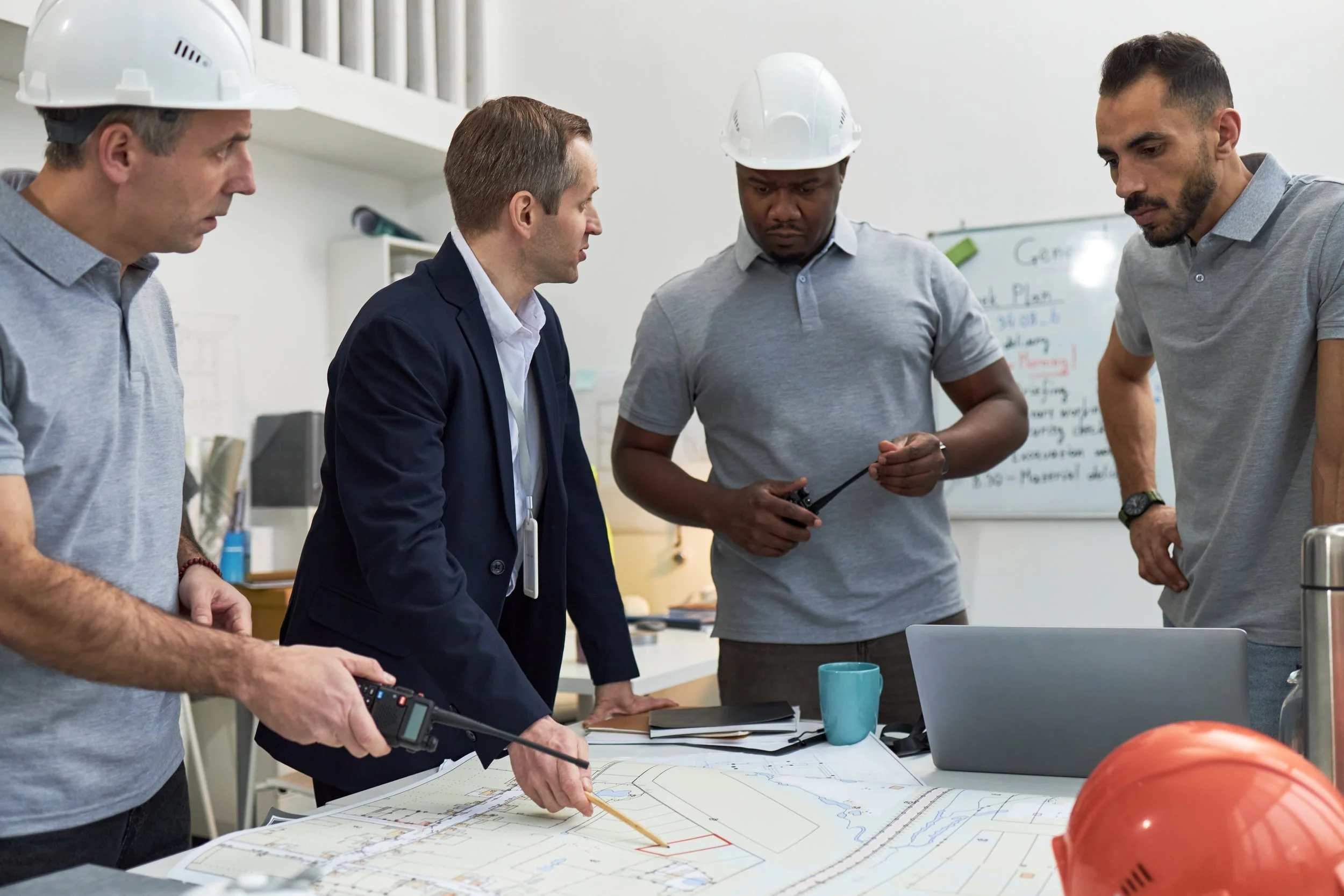 Group of four men, some wearing construction helmets, examining architectural plans and discussing a construction project in an office or construction site.