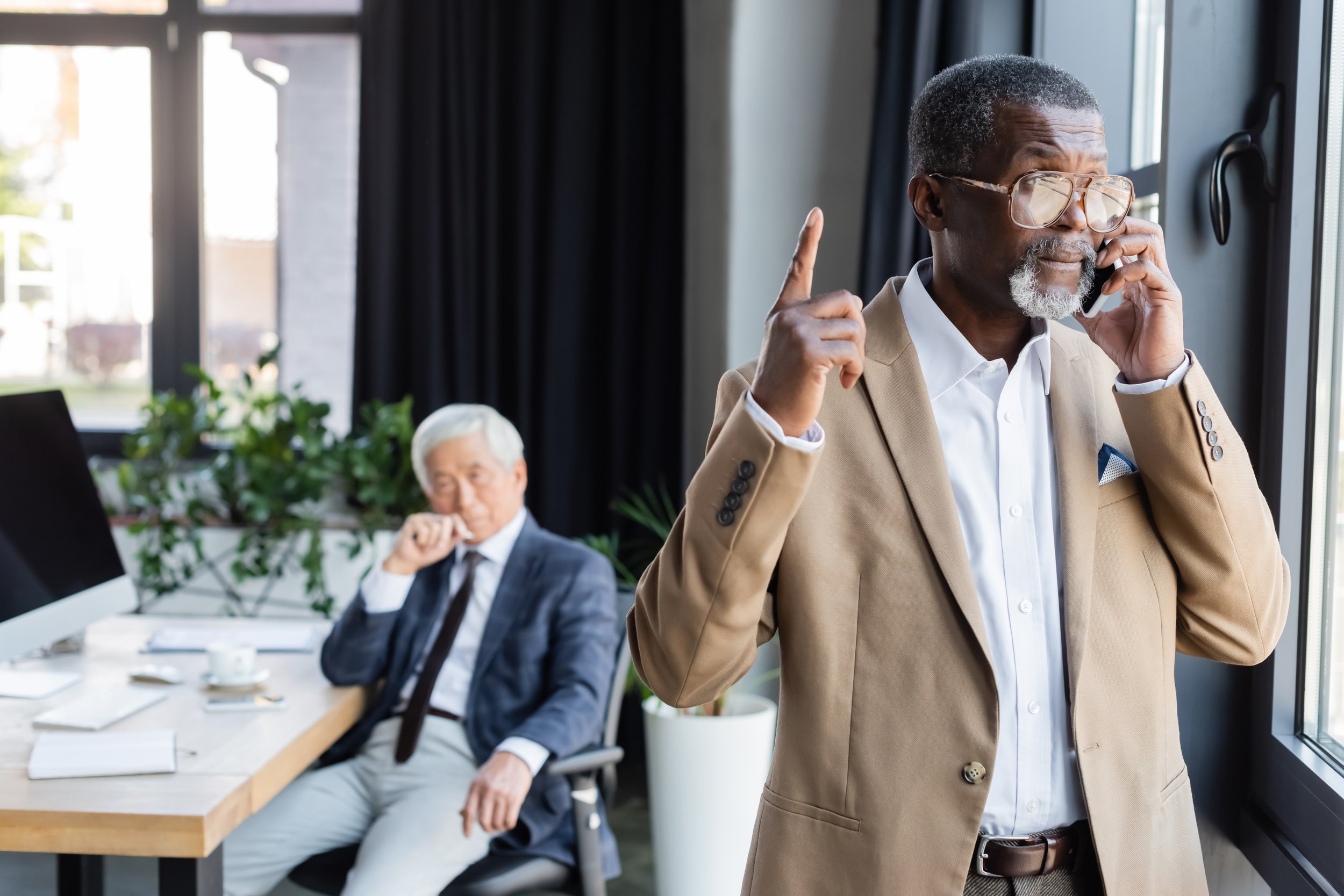 An older man with glasses and a beard, dressed in a beige blazer and white shirt, is standing and talking on the phone near a large window. In the background, another man with white hair, dressed in dark clothing, is sitting at a conference table with glasses, a cups, and notebooks, appearing to be listening or thinking.