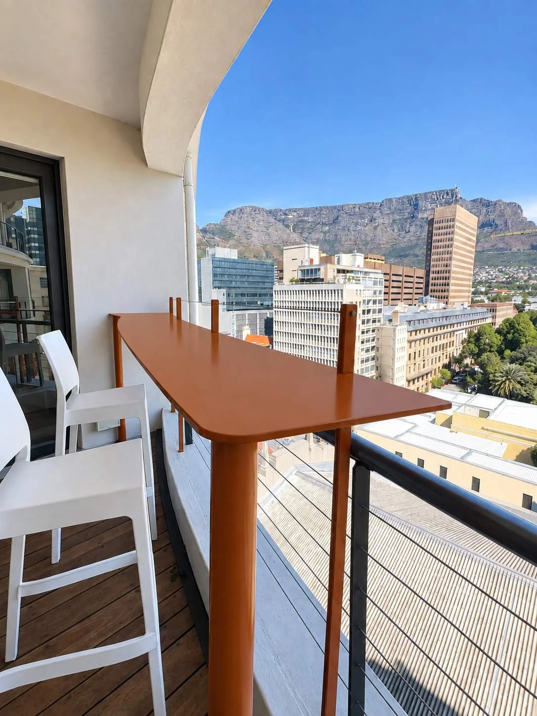 Terracotta powder-coated steel balcony bar table with white bar stools and a view of Table Mountain and Cape Town