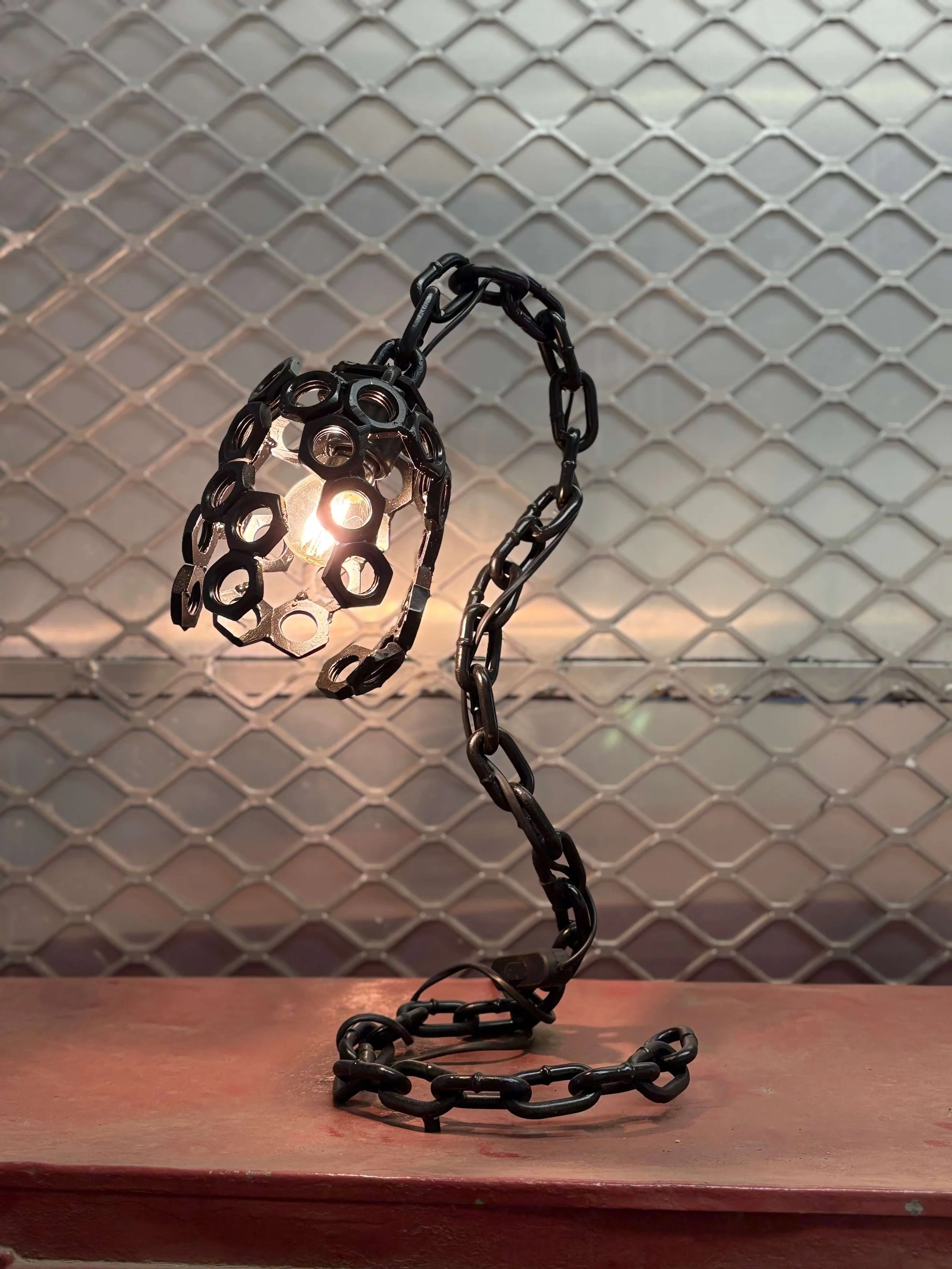 A unique table side lamp made of black metal chain links and metal washers. The fixture has a bulb emitting warm light.