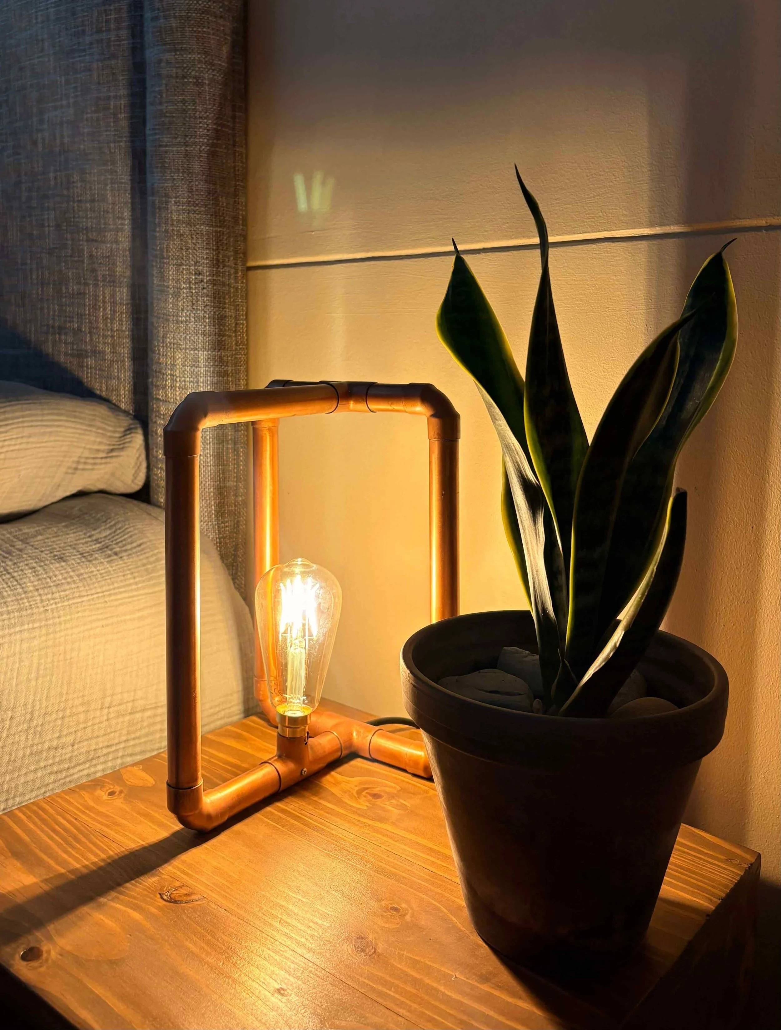 A modular polished copper lamp next to a potted plant on a wooden bedside table.