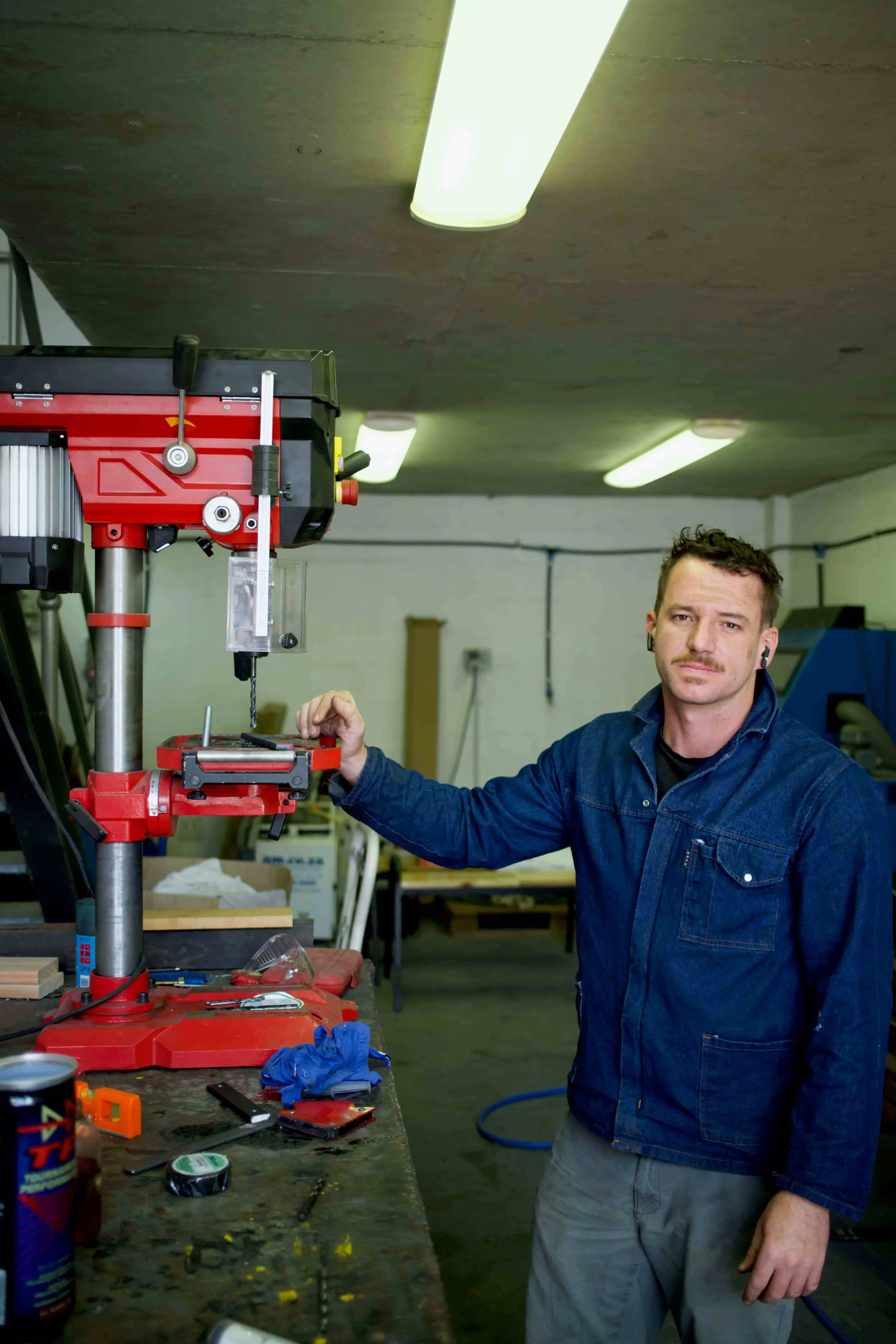 A man working in a workshop, operating a red drill press with his left hand, wearing a denim shirt and gray pants. The workshop has various tools and supplies on a cluttered workbench, with fluorescent ceiling lights overhead.