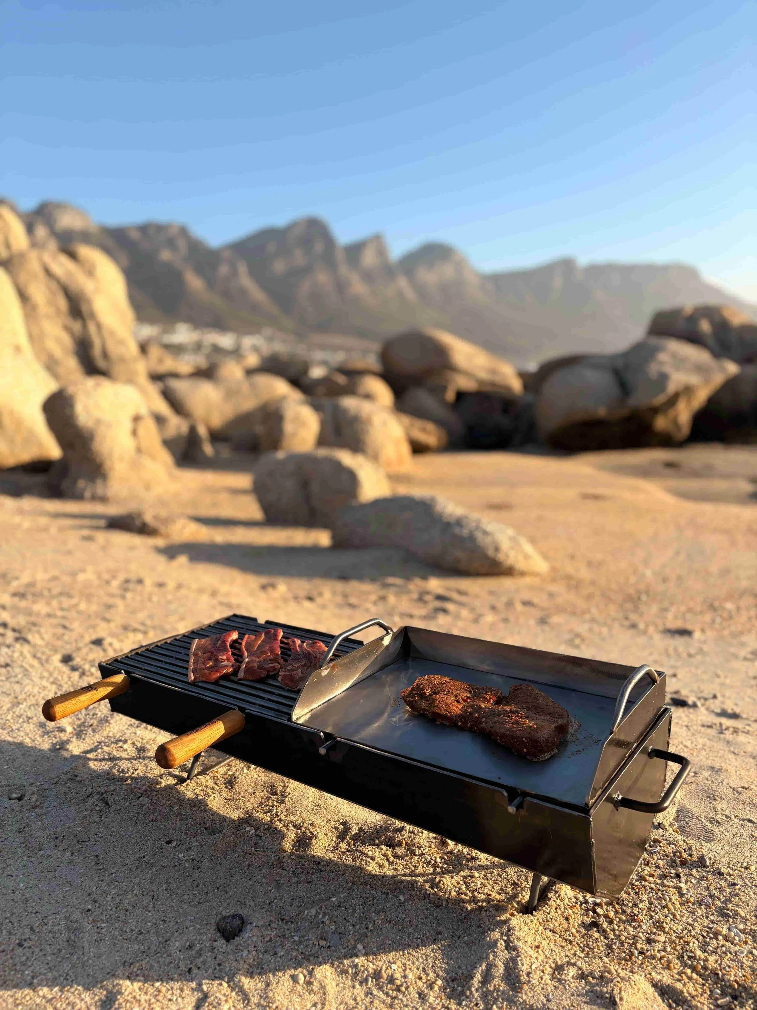 A portable braai sitting on a sandy beach cooking pork chops and a piece of steak, with blurred rocks and mountains in the background under a clear blue sky.