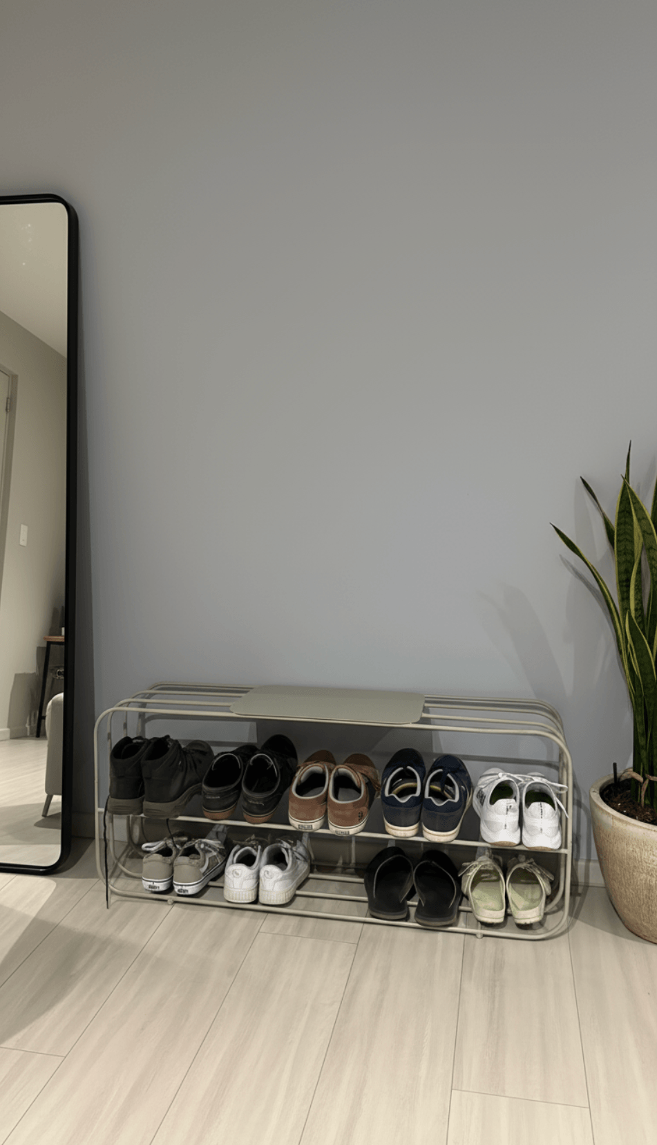 A minimalist shoe rack with two shelves containing sneakers and boots, adjacent to a full-length mirror and a potted plant against a plain gray wall.