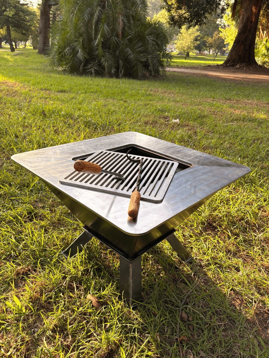 A steel braai that is used as a fire pit with flames burning, situated outdoors on a grassy area with a house and deck in the background, overlooking a scenic landscape at sunset.
