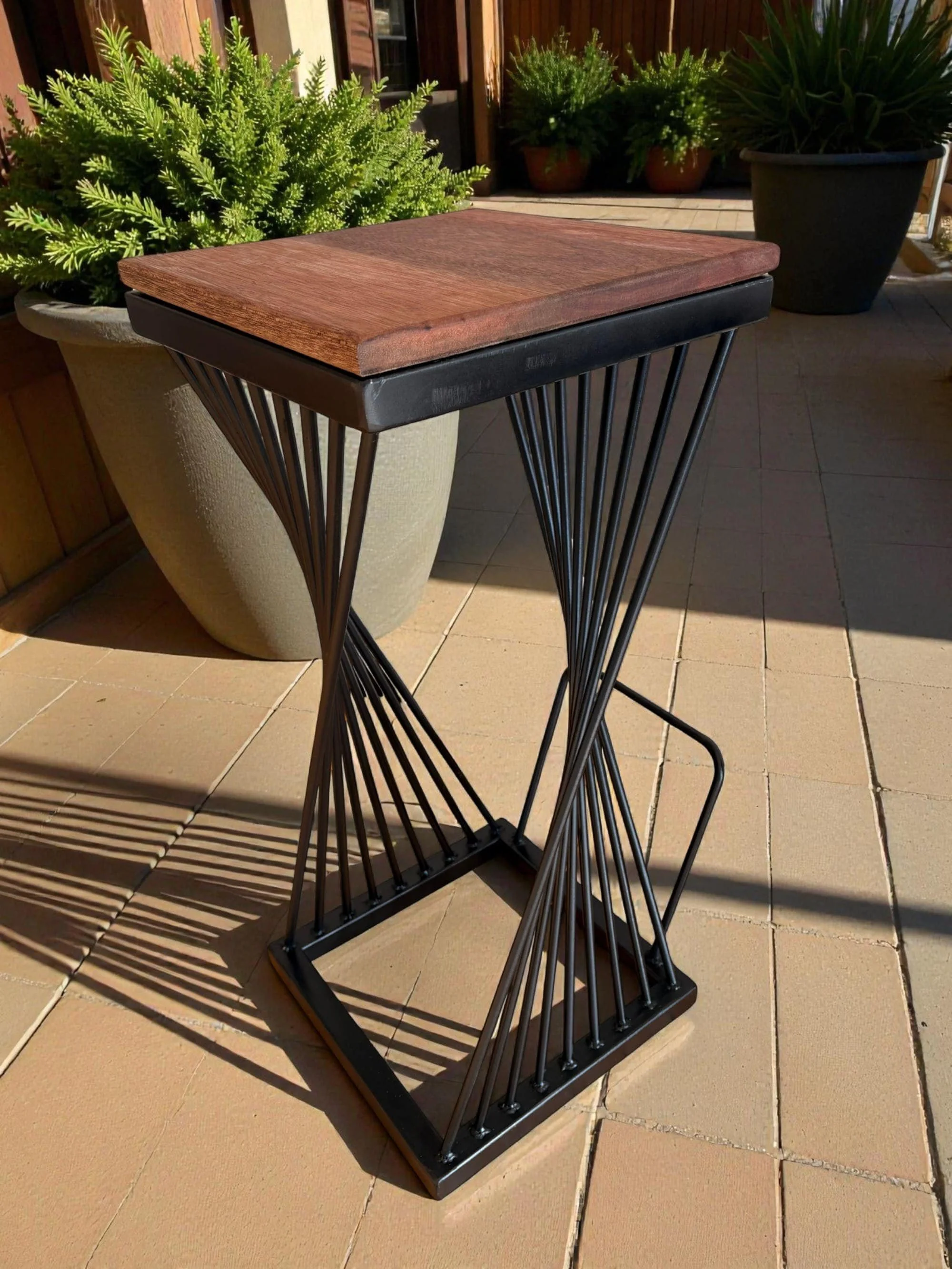 A industrial bar stool with wood seat and steel frame on a sunlit outdoor patio, with potted green plants in the background.