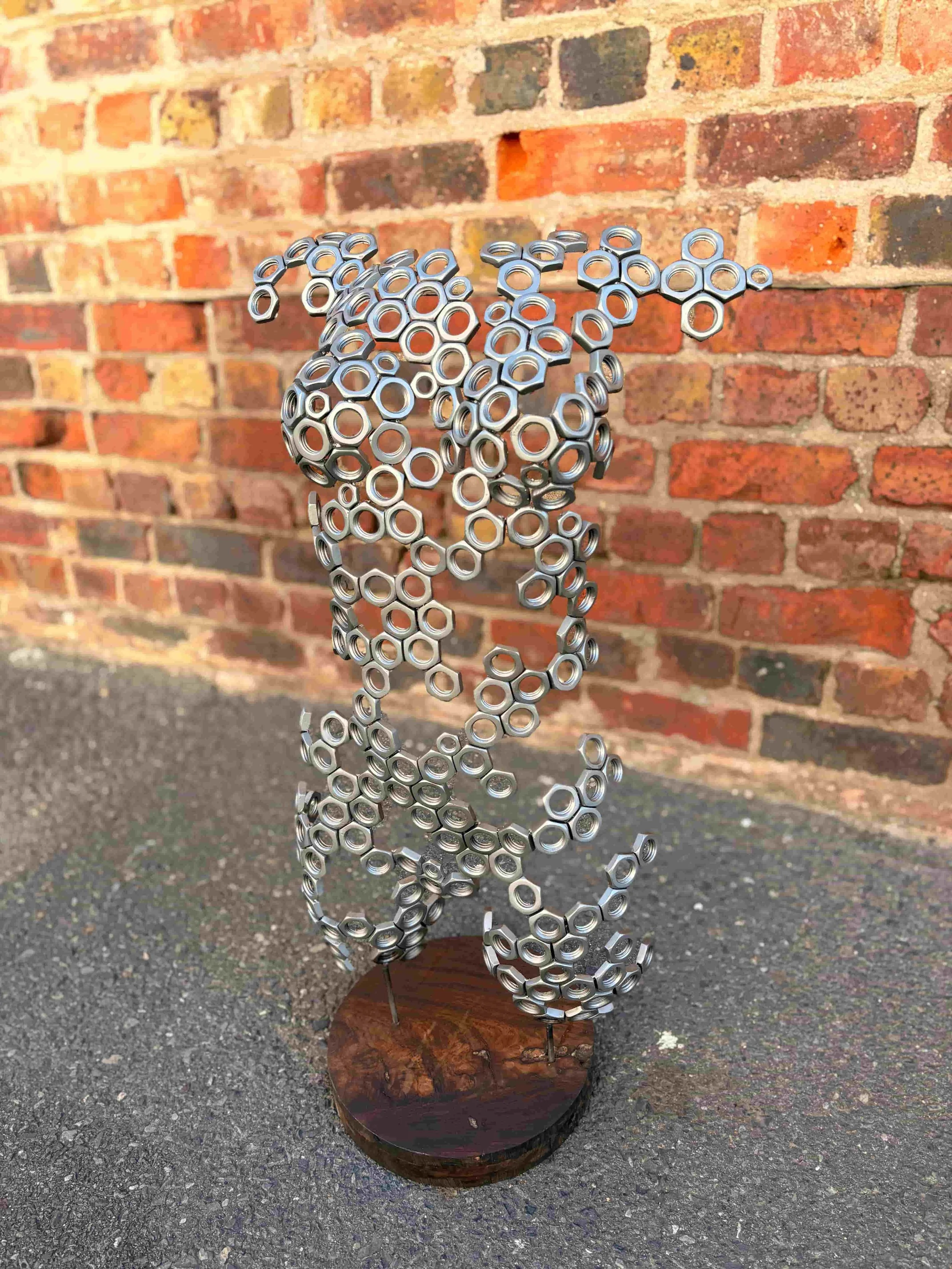 A sculpture made from metal nuts assembled in the shape of womans torso, mounted on a wooden base, placed outdoors on asphalt with a red brick wall in the background.