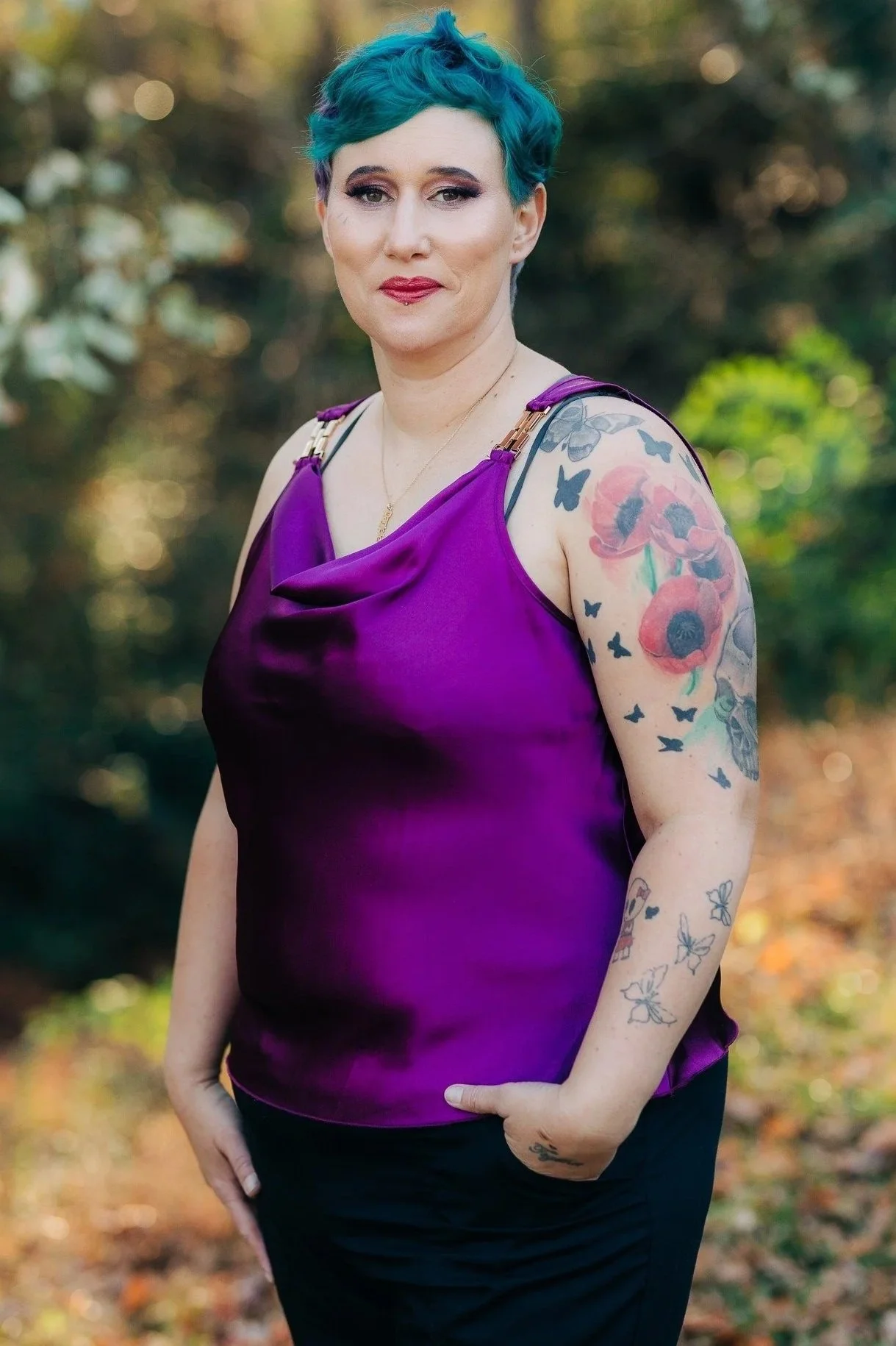Woman with blue and purple short hair wearing a purple satin top and black pants in an outdoor setting with trees and sunlight in the background. Danielle Torres, business owner at Rustic Roots Studio LLC, in Altavista, VA.