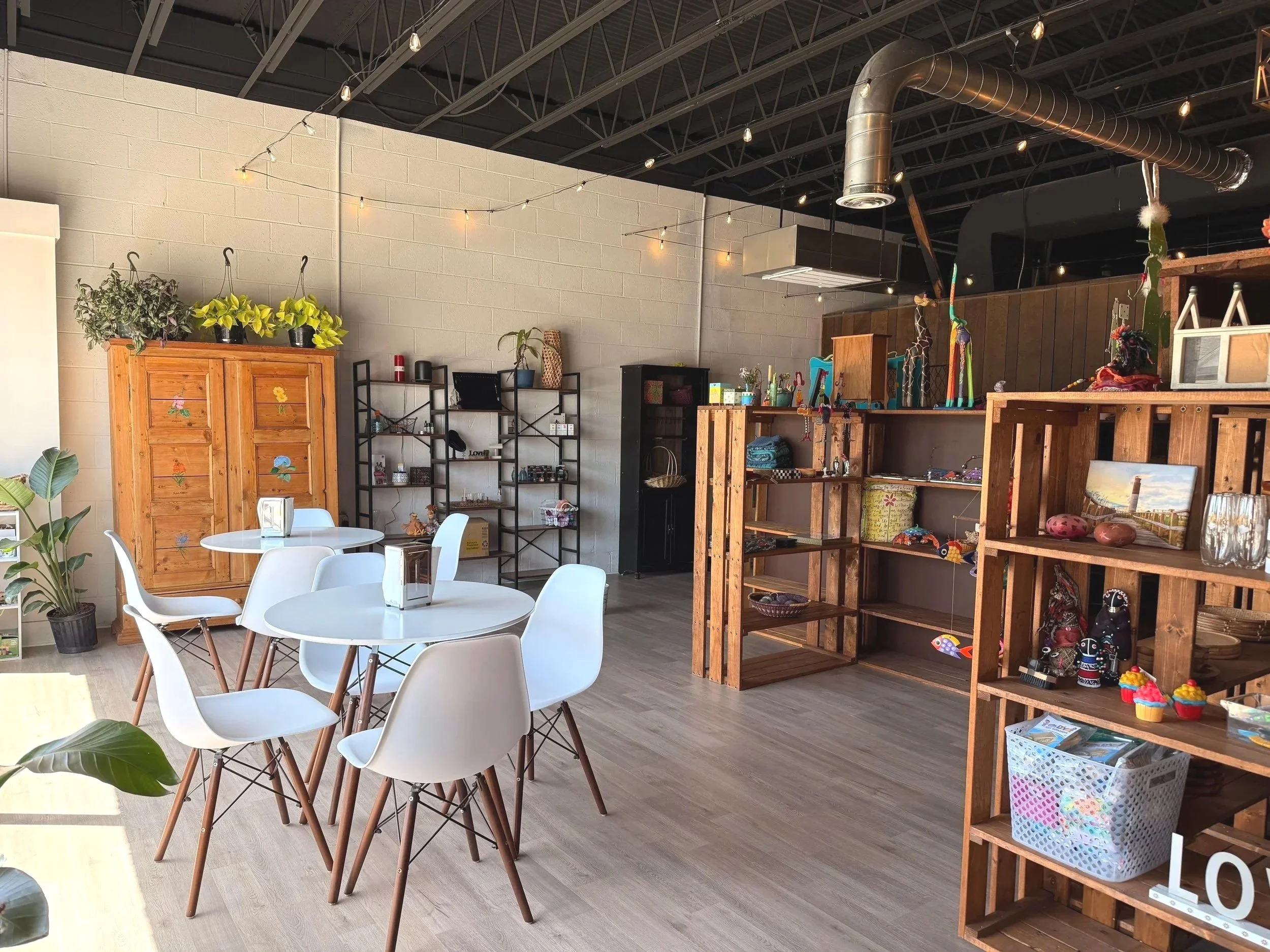 Interior of a store or cafe with white round tables and white chairs, wooden shelves with decorative items, plants, and string lights on the ceiling.