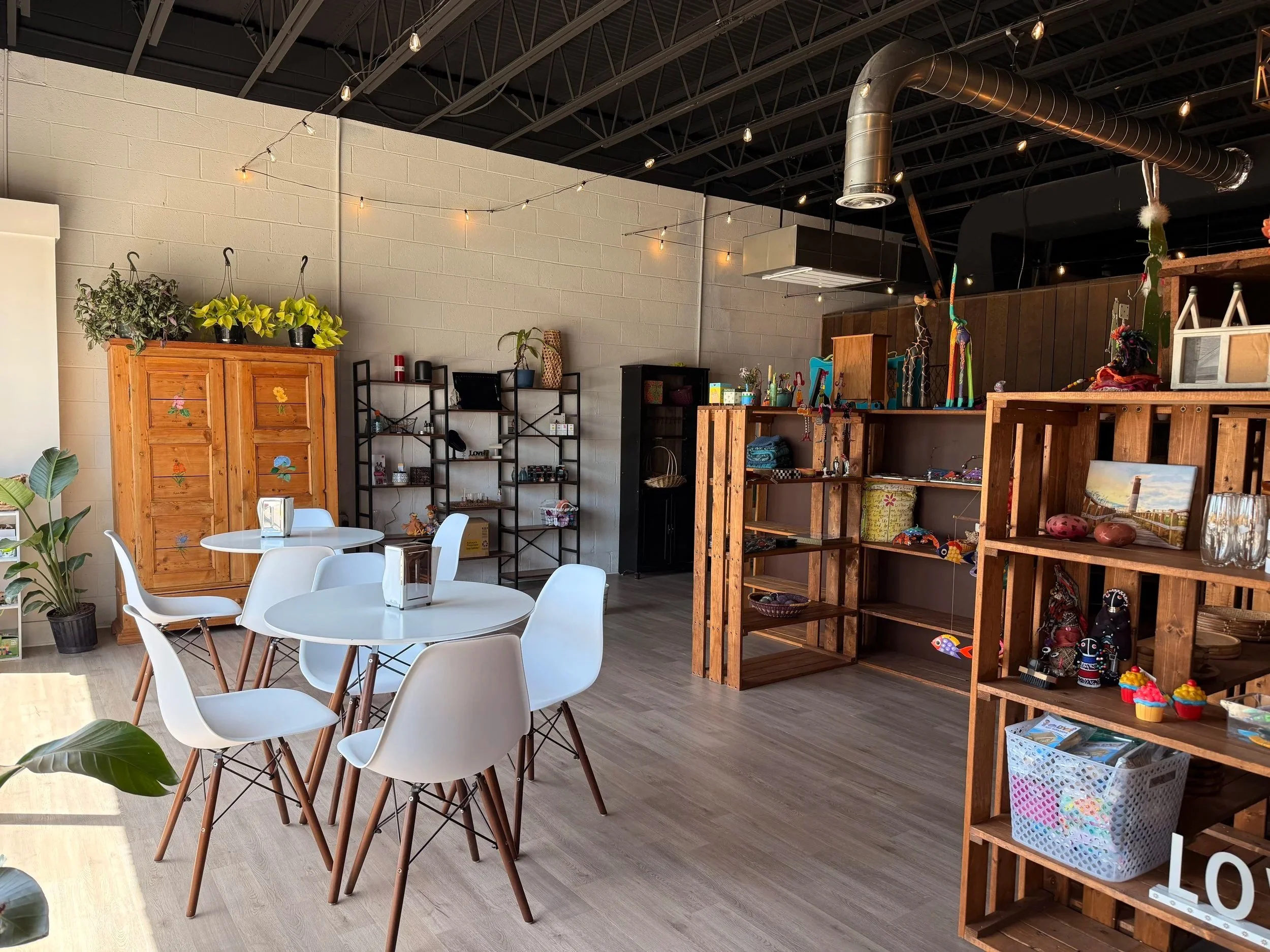 Interior of a store with wooden and metal shelves displaying various decorative items, plants, and small toys, with a high exposed ceiling and string lights.