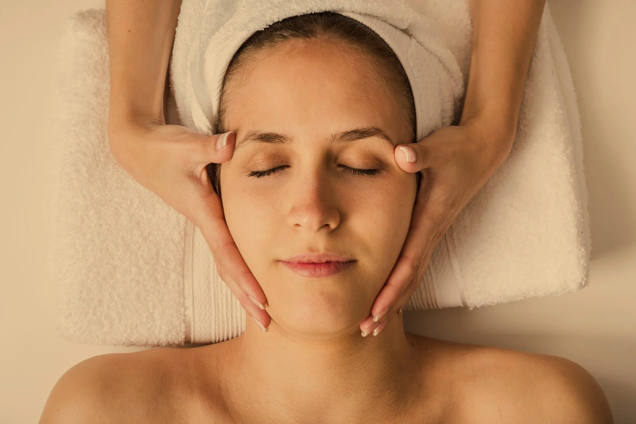 A person lying face up on a massage table with eyes closed, receiving a facial massage from a therapist. The therapist's hands gently cradle the person's face and neck.