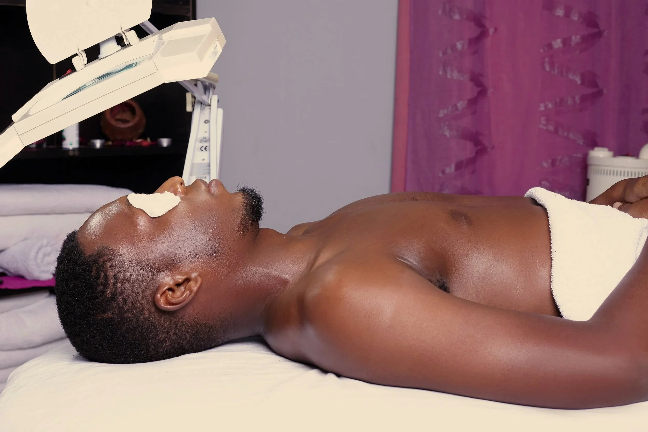 A person lying on an examination table during a skincare or facial treatment, with a facial mask applied and an esthetician or technician attending to them.