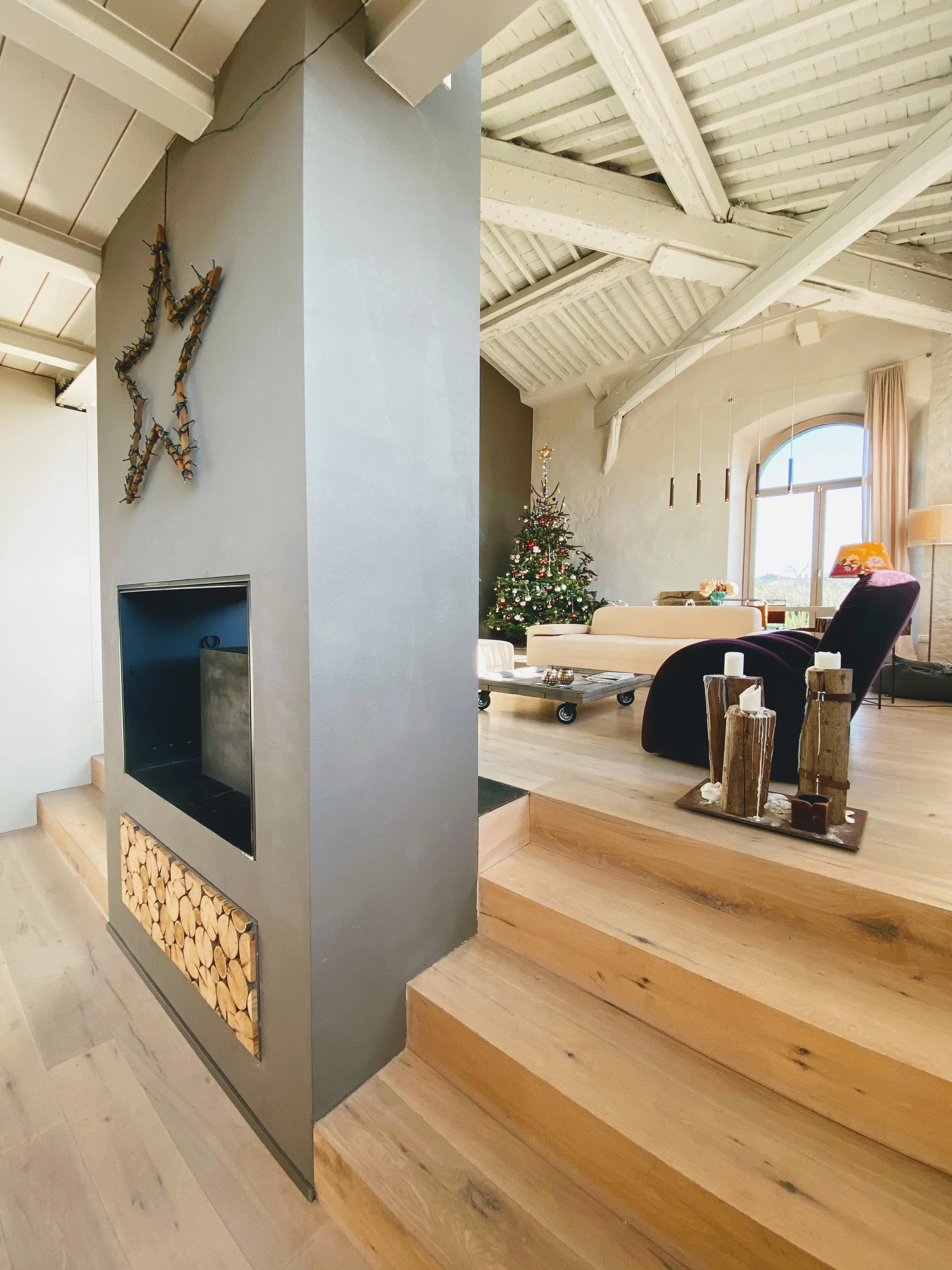 Interno di una casa con soffitto a travi bianche, un albero di Natale decorato vicino a un divano. Decorazioni natalizie su un tavolo e un camino in cartongesso con un mucchio di legna sopra. Illuminazione naturale dalla finestra.