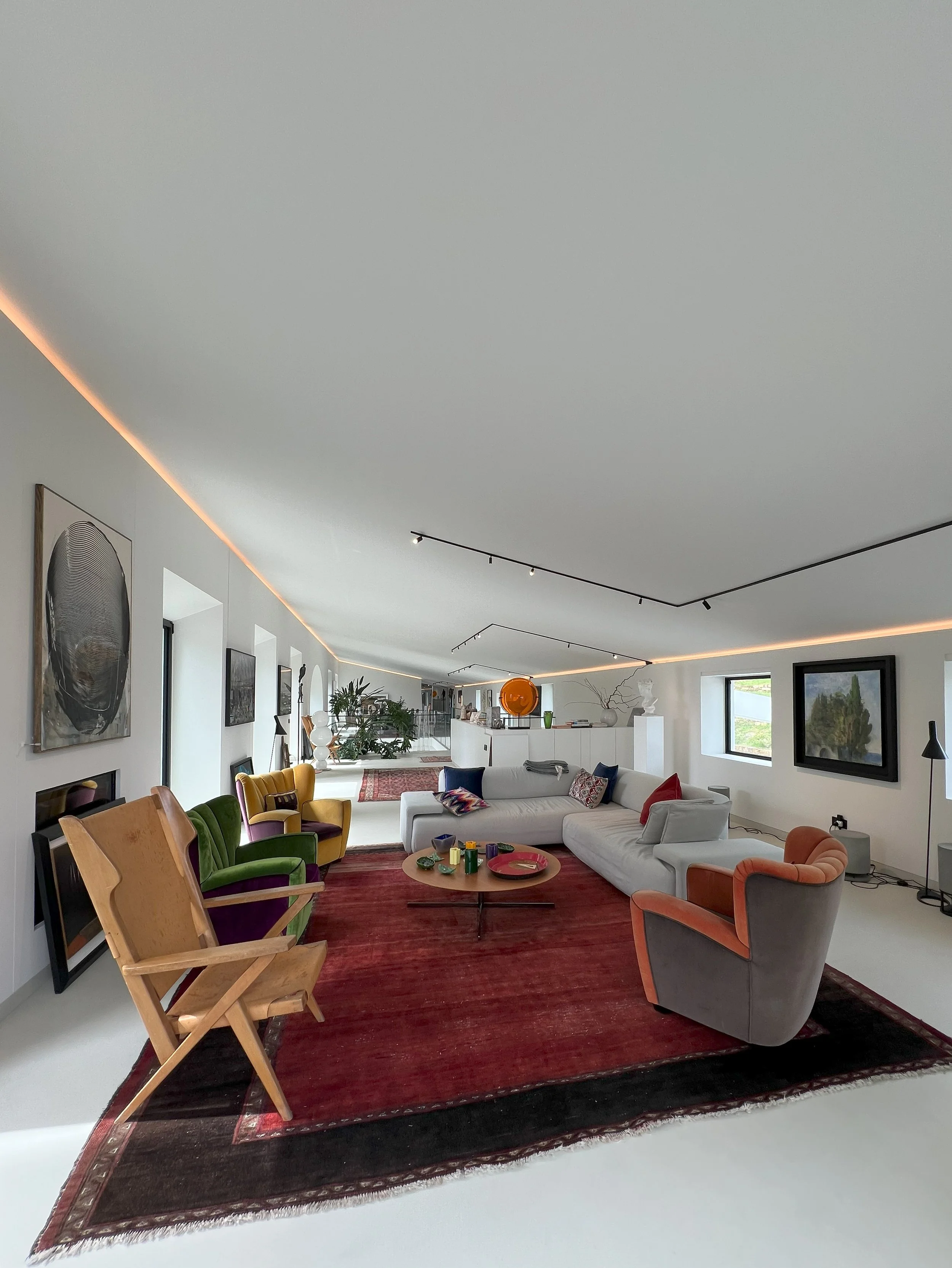 Soggiorno moderno con divano bianco, poltrone colorate, tappeto rosso e pareti decorate con quadri e arte, grandi finestre e un soffitto bianco lineare.