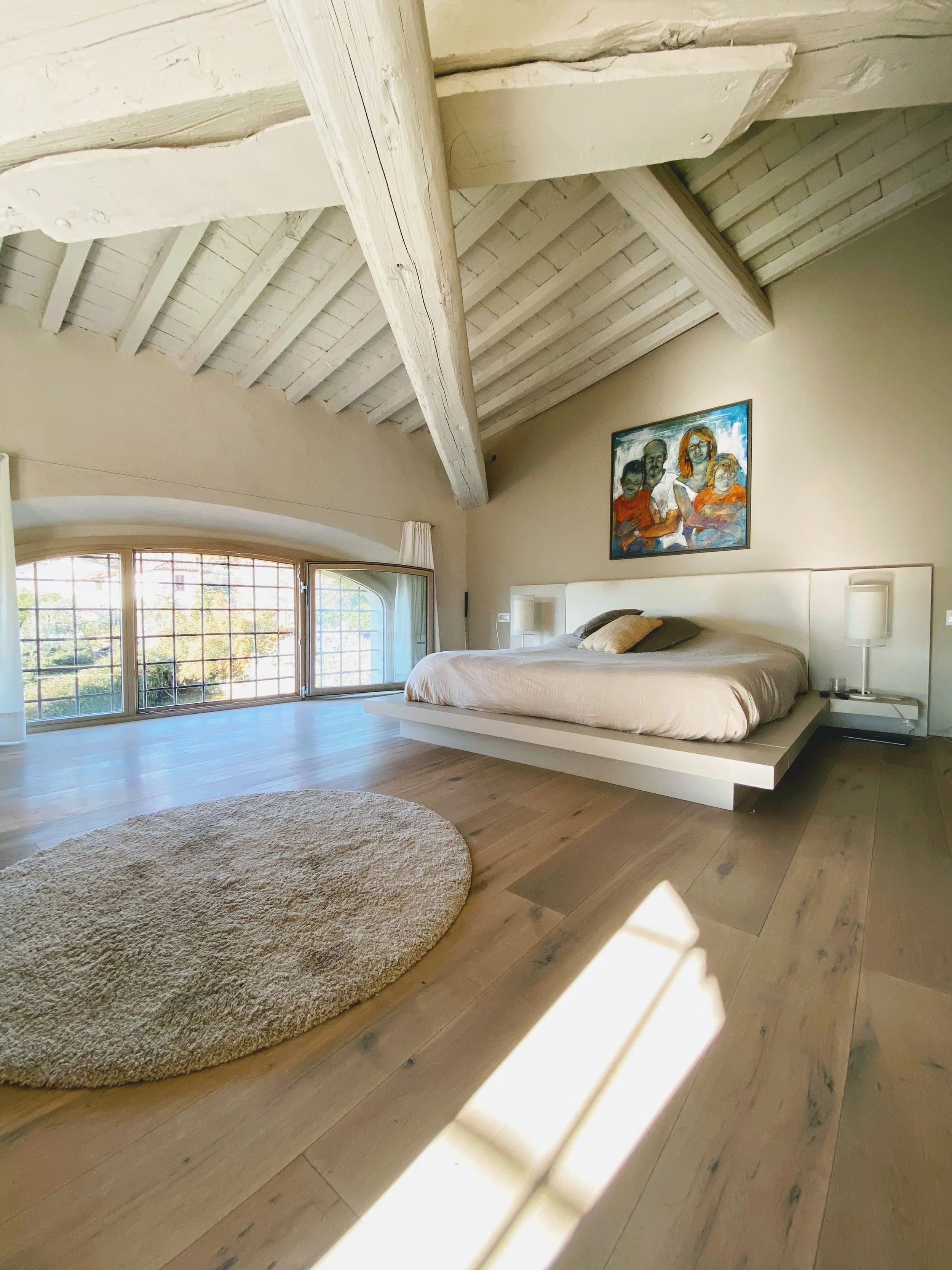 Camera da letto con letto grande, arredamento minimalista, pavimento in legno chiaro, pareti beige, una finestra grande open space con tende bianche e quadro colorato sopra il letto, soffitto con travi in legno bianco, tappeto rotondo beige.