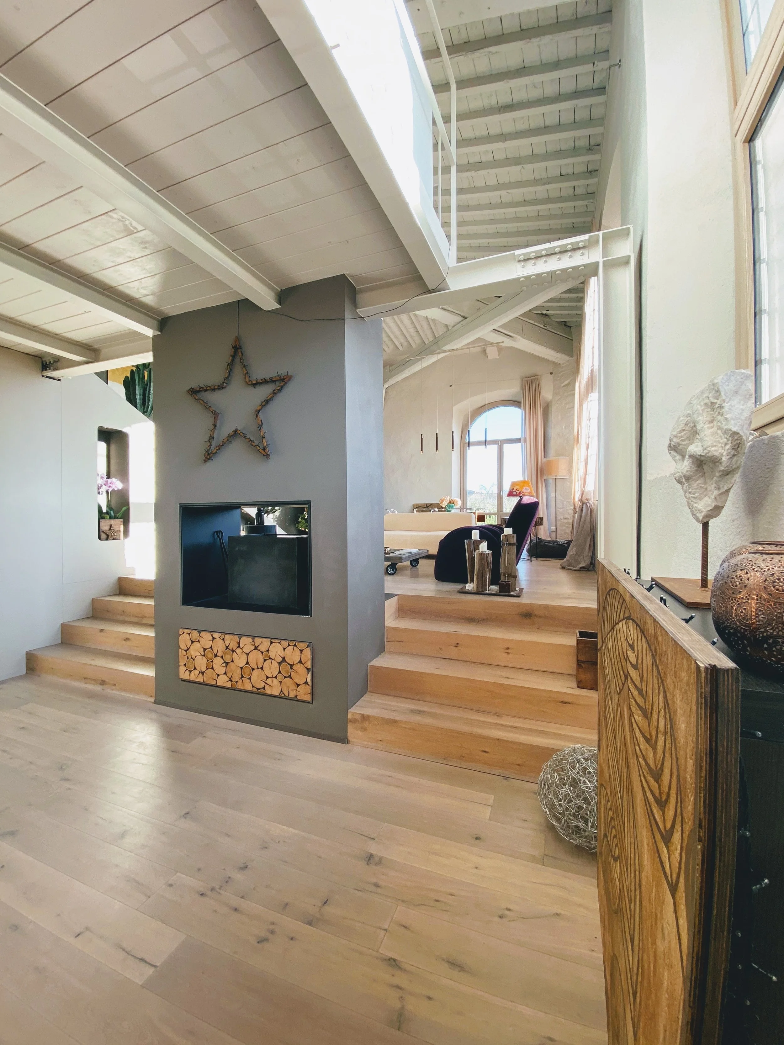 Interno di una casa moderna con area soggiorno al piano superiore, parete con decorazione a stella e camino, sedia a dondolo, oggetti decorativi in legno e pietra, grande finestra con tende chiare.