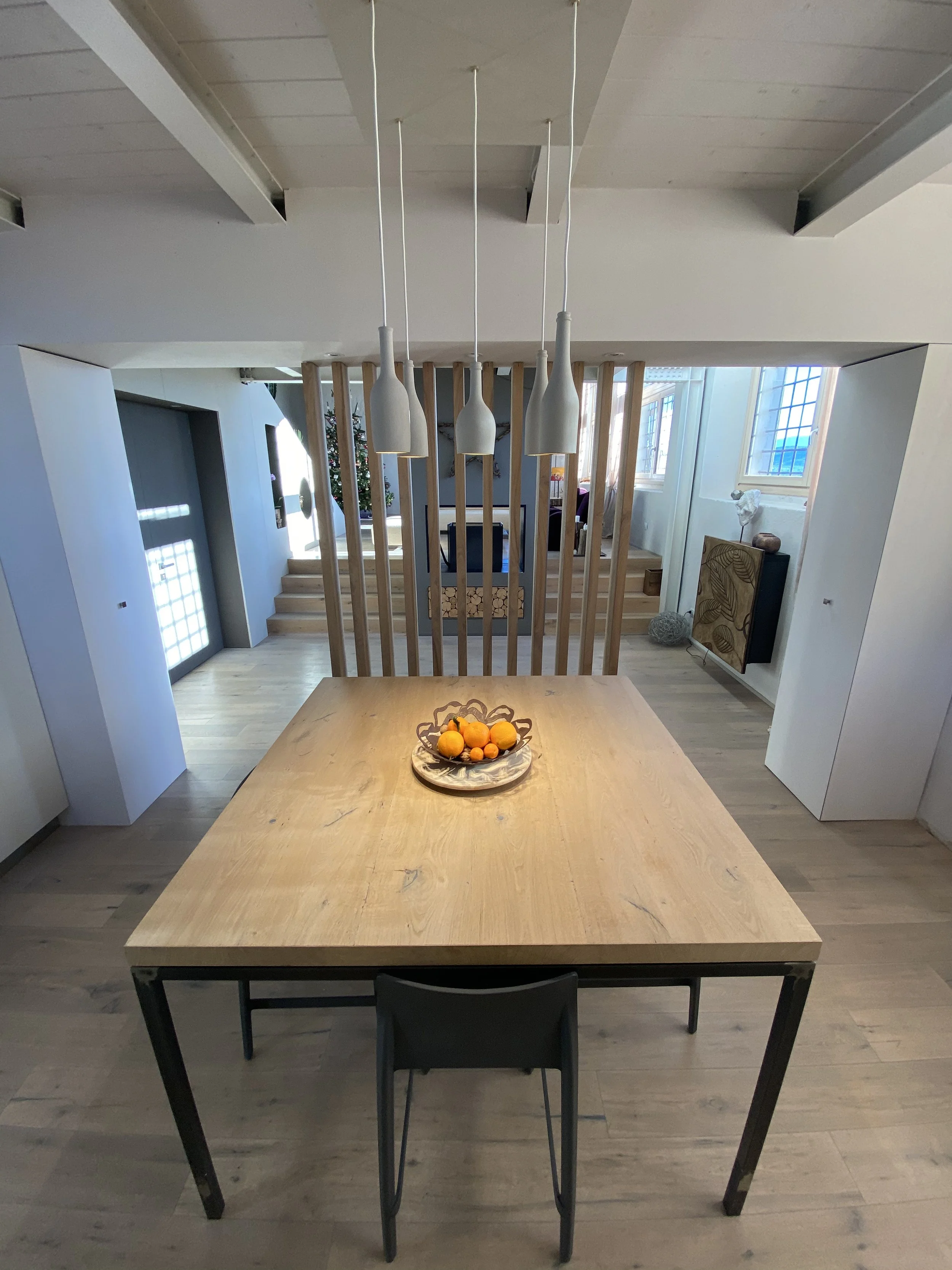 Una stanza moderna con un tavolo di legno al centro, decorato con una ciotola di arance. Sopra il tavolo ci sono quattro lampade sospese bianche. Sullo sfondo, una zona salotto con un divano e una scala in legno che porta a un piano superiore. Finest