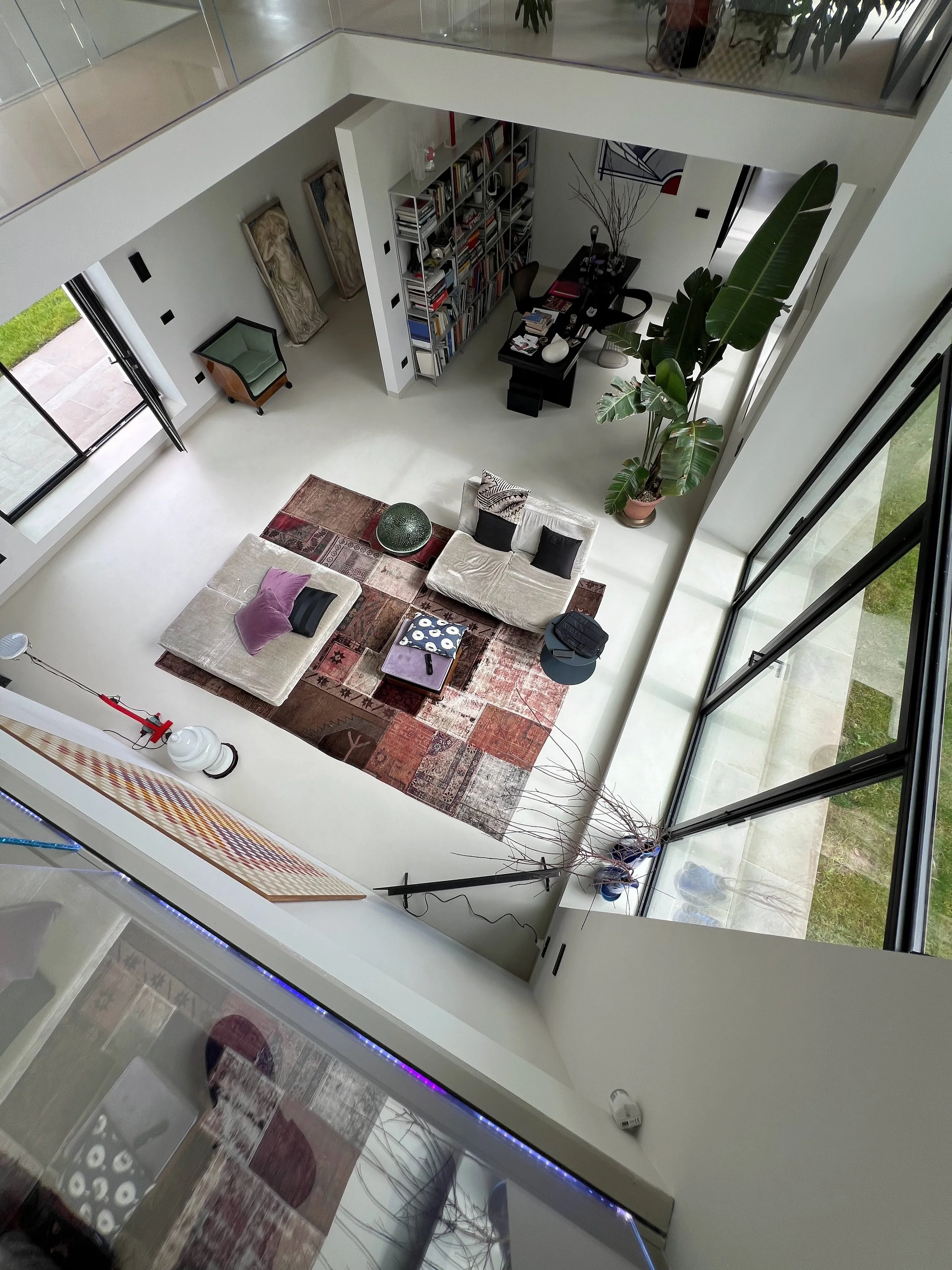 Vista dall'alto di un soggiorno moderno con grandi finestre, divani, tappeto colorato, piante grandi e una libreria