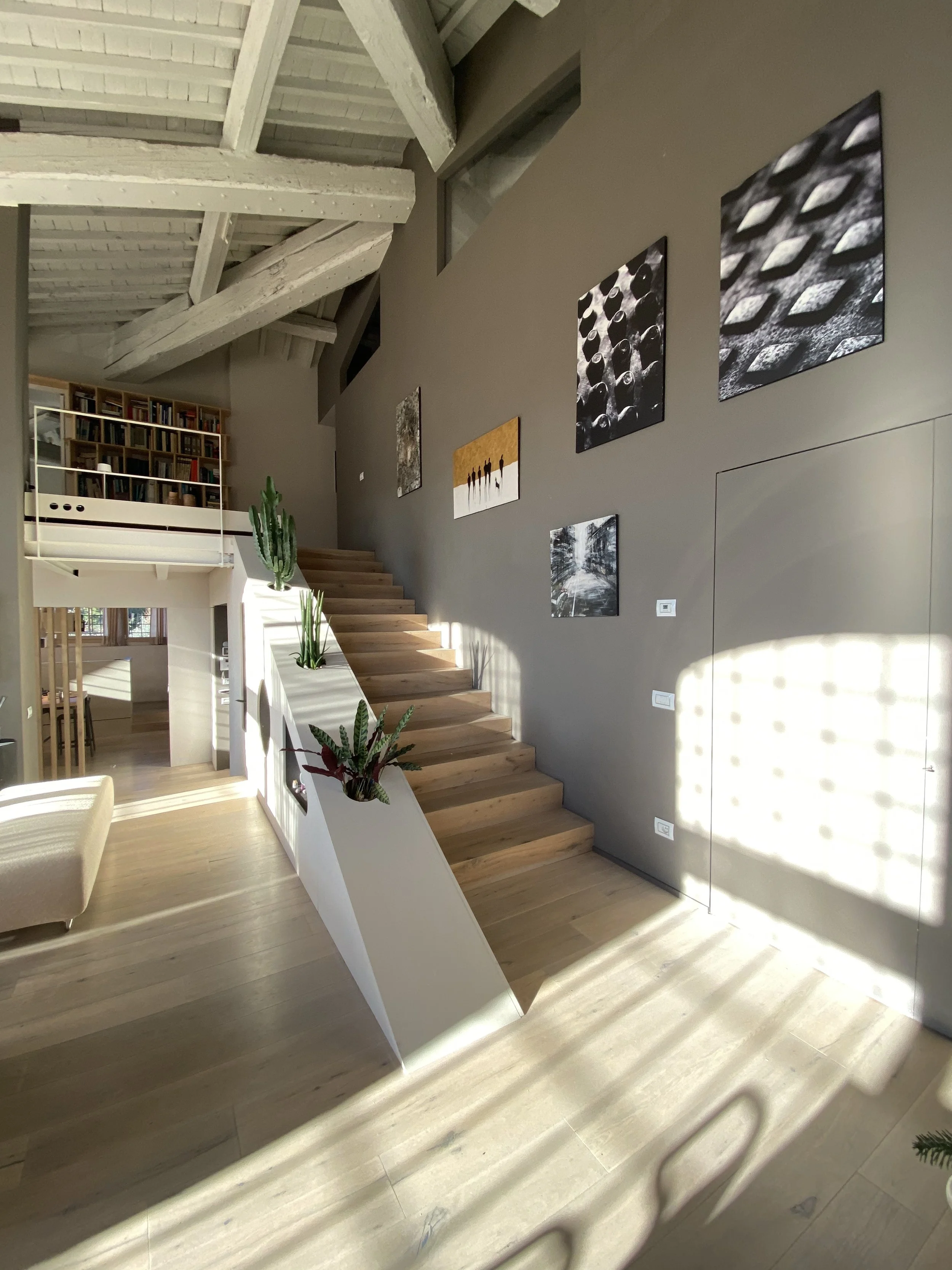 Interno di una casa con parete grigia decorata con quadri, scala in legno, piante verdi e luce naturale che crea ombre sul pavimento.