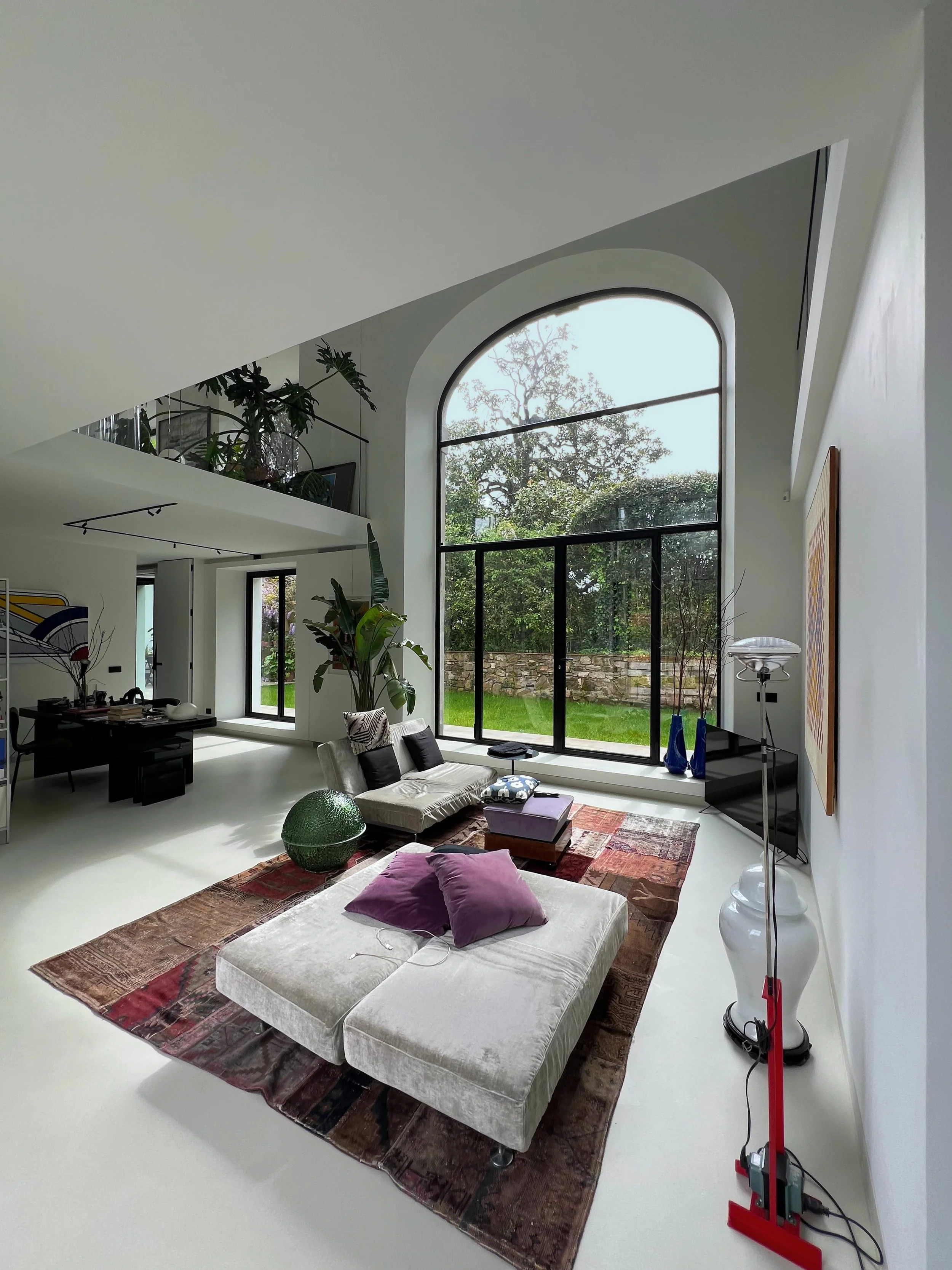 Interno di un soggiorno moderno con grandi finestre ad arco, divano beige con cuscini viola, tappeto colorato, piante verdi, lampada a stelo bianca, TV, e decorazioni vari.