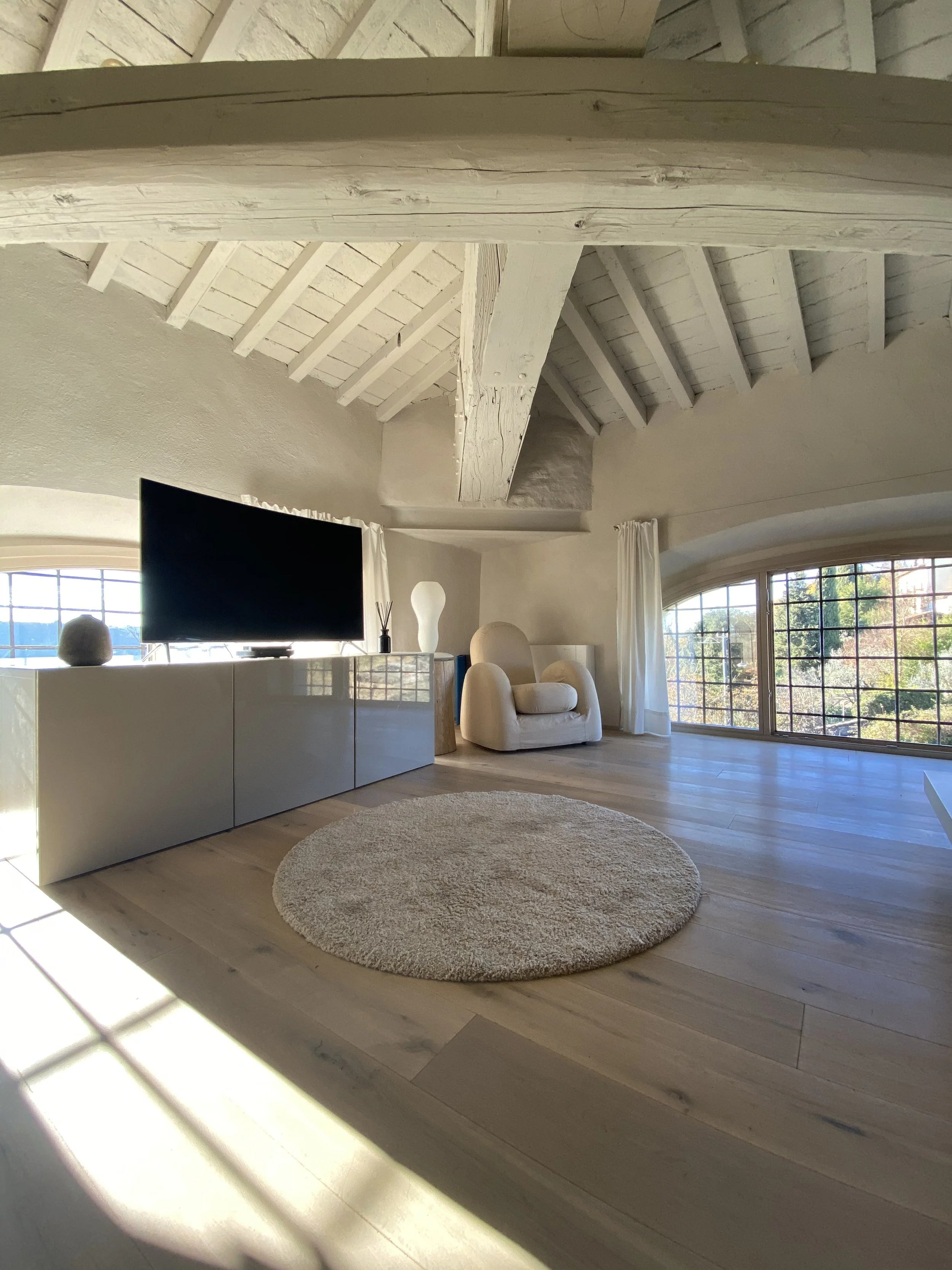 Interno di un soggiorno con soffitto con travi di legno bianco, grande finestra con vista su un paesaggio naturale, poltrona beige, mobile TV bianco lucido, tappeto rotondo beige, decorazioni minimaliste, luce naturale.