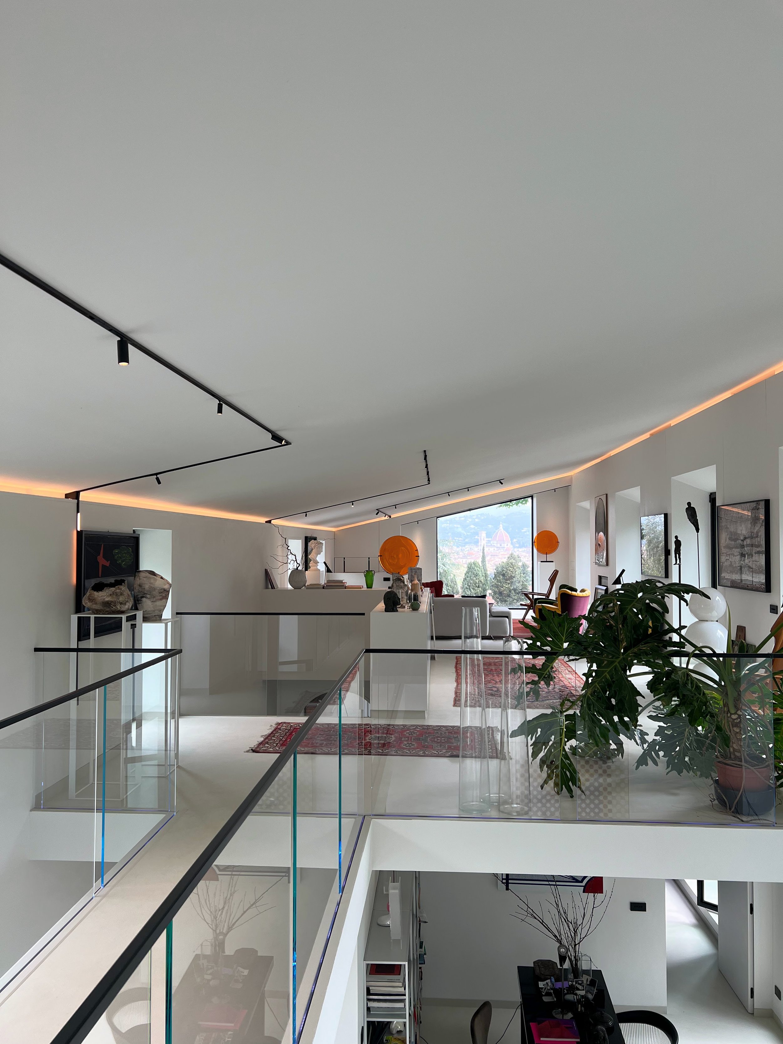 Interno di un appartamento moderno con spazio aperto, diviso tra una zona living con divani e decorazioni artistiche, e una zona superiore con piante e abbellimenti, grandi finestre che offrono vista sulla città.
