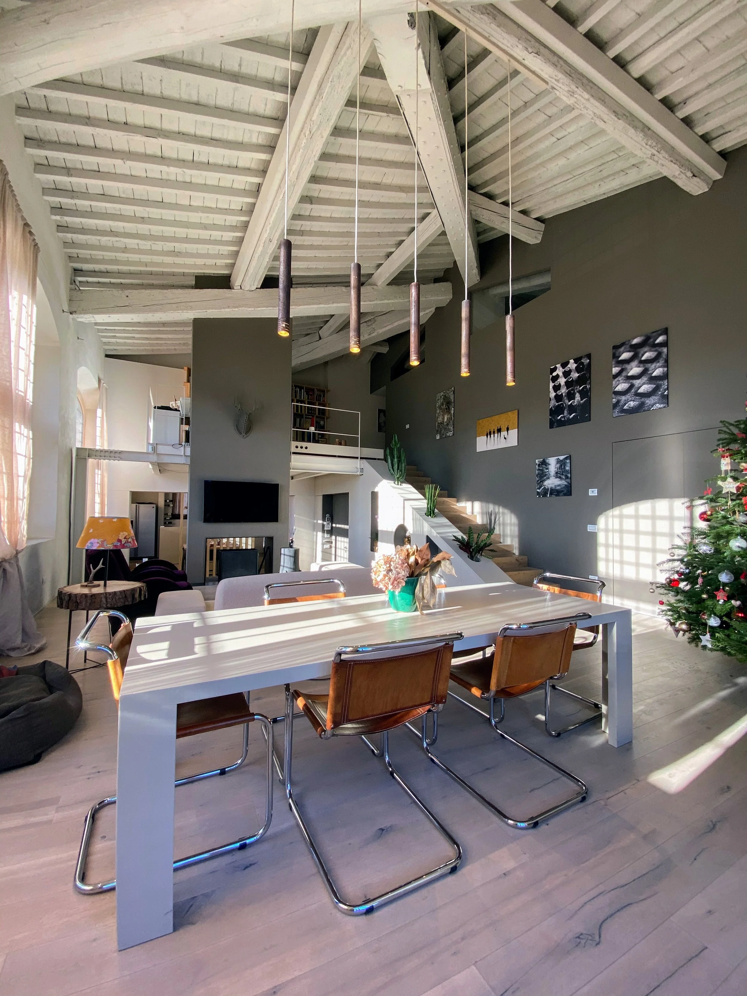 Interno di una casa con soffitto a spina di pesce e travi in legno bianco, grande tavolo bianco con sedie in pelle marrone, decorazioni, al centro un vaso con fiori secchi, decorazioni su pareti.