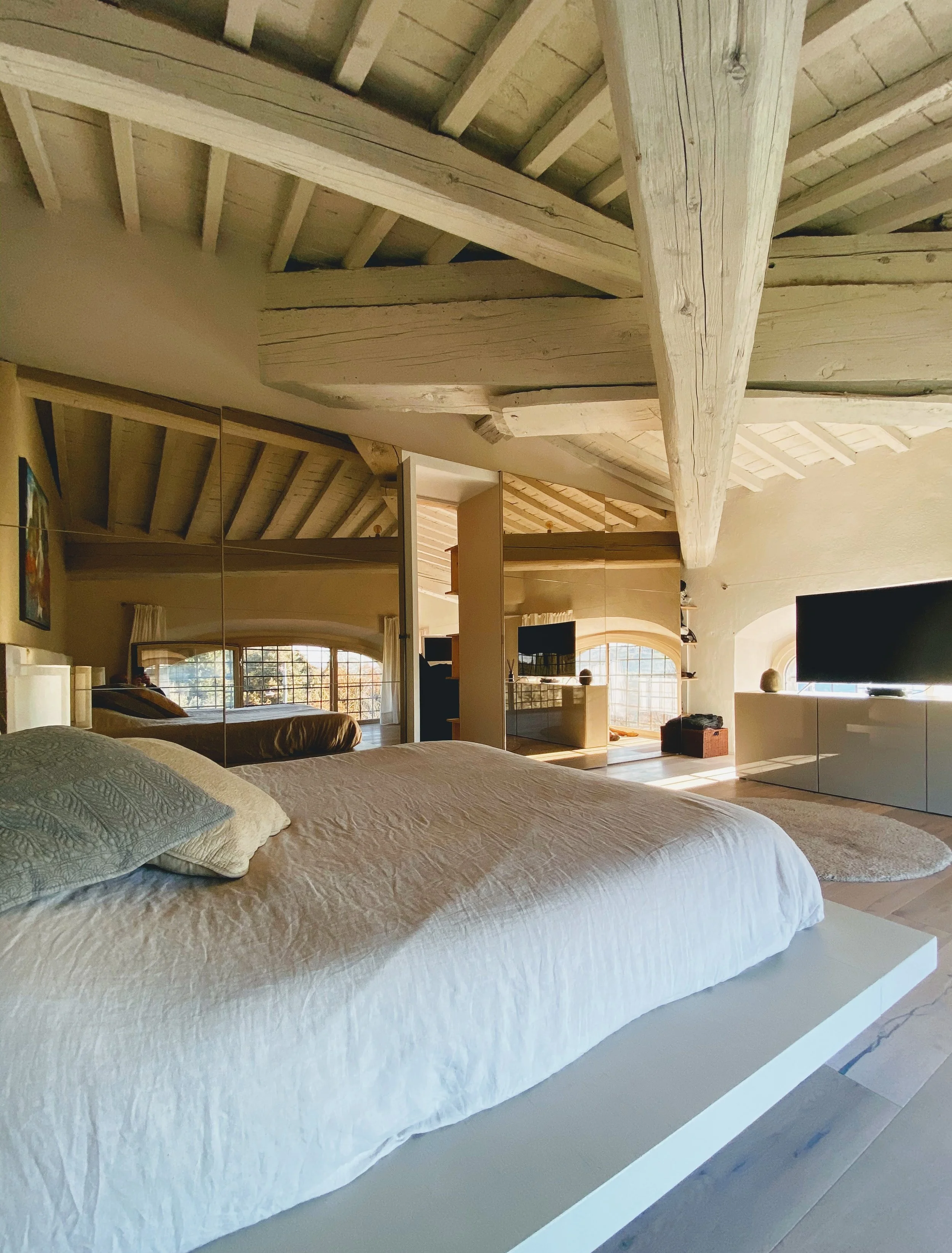 Camera da letto con un letto matrimoniale con cuscini, pareti con specchi, finestre con tende, e un televisore, con soffitto a vista in legno.