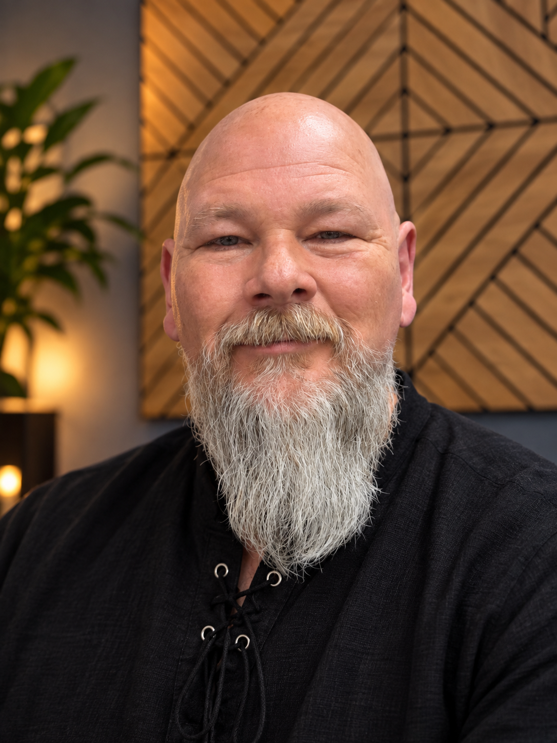 Close-up of a smiling bald man with a full white beard, wearing a black shirt, in a warmly lit room with wooden wall art and a plant in the background.