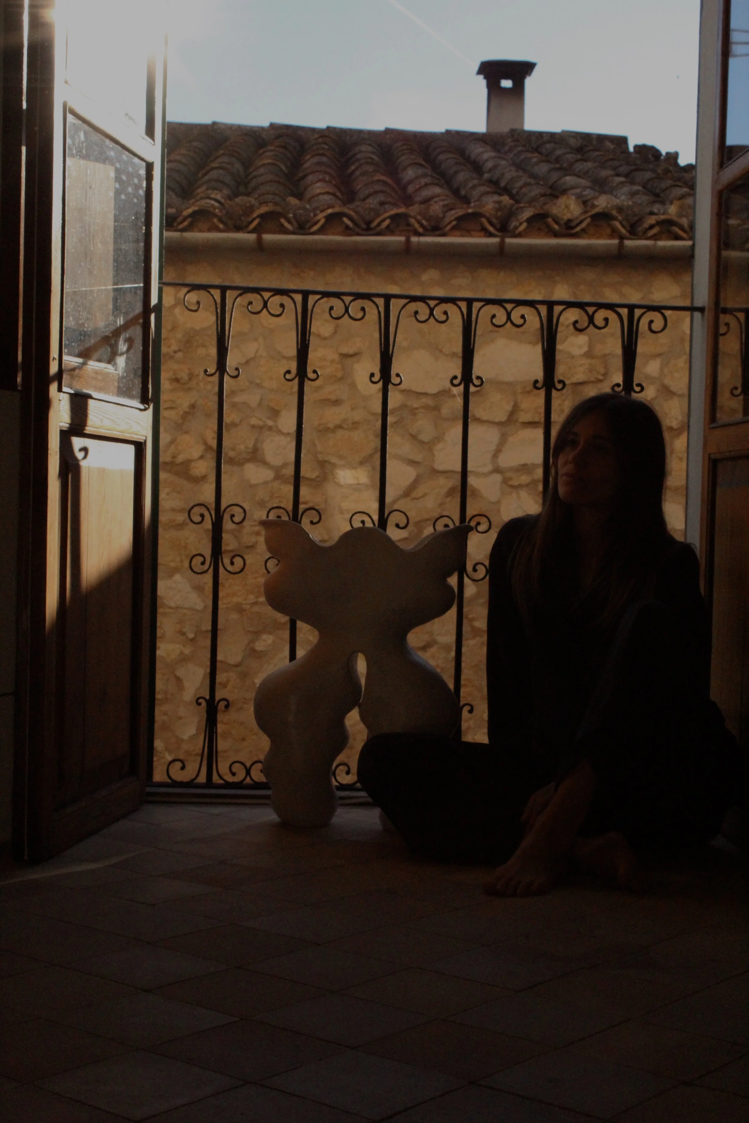 Silhouette of a woman sitting on the floor near a decorative sculpture on an indoor balcony with open wooden windows, stone wall background, and tiled floor.