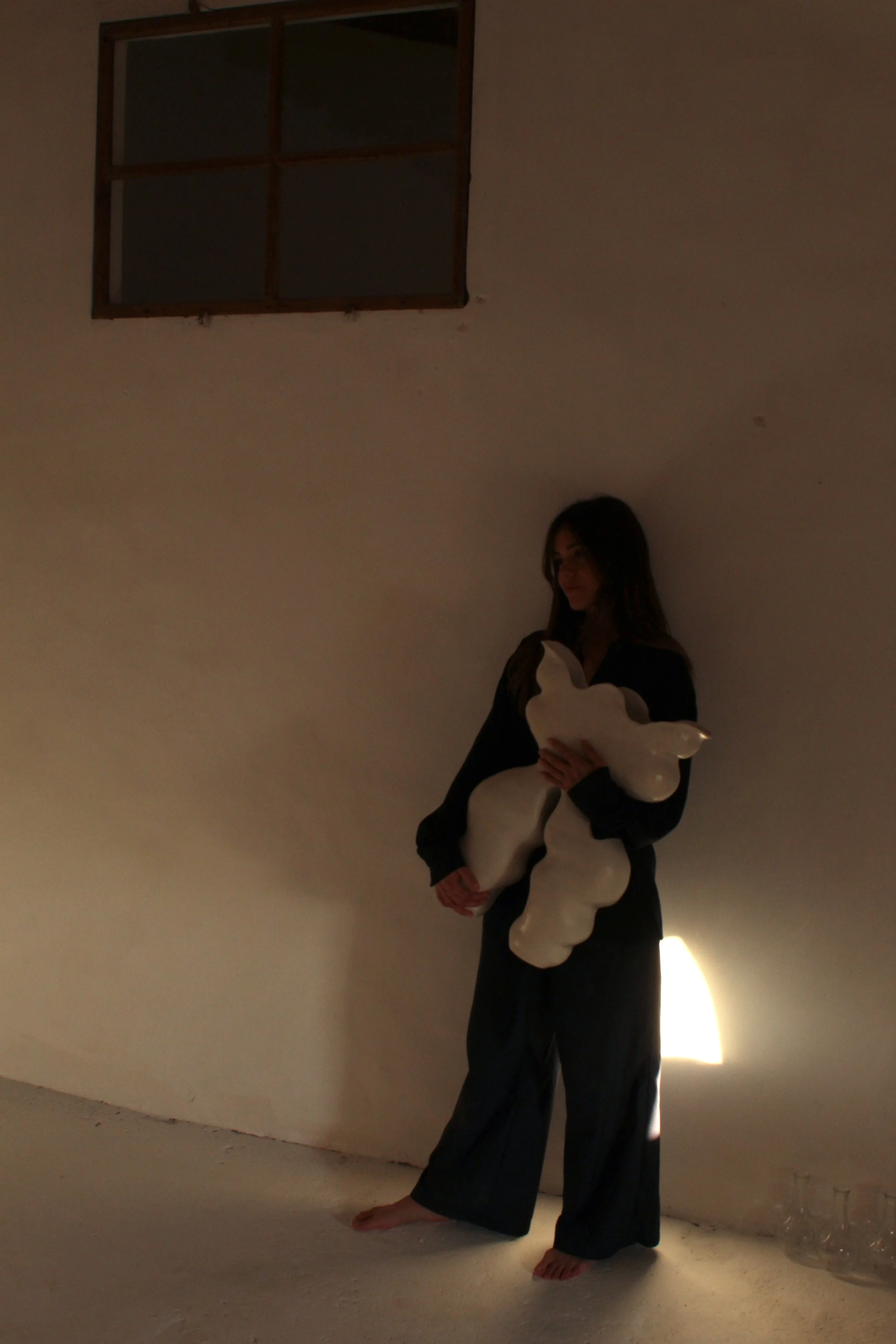 A woman standing barefoot against a plain wall, holding a white sculpture with a cloud-like shape. There is a small window near the ceiling and a light source illuminating the lower right corner.