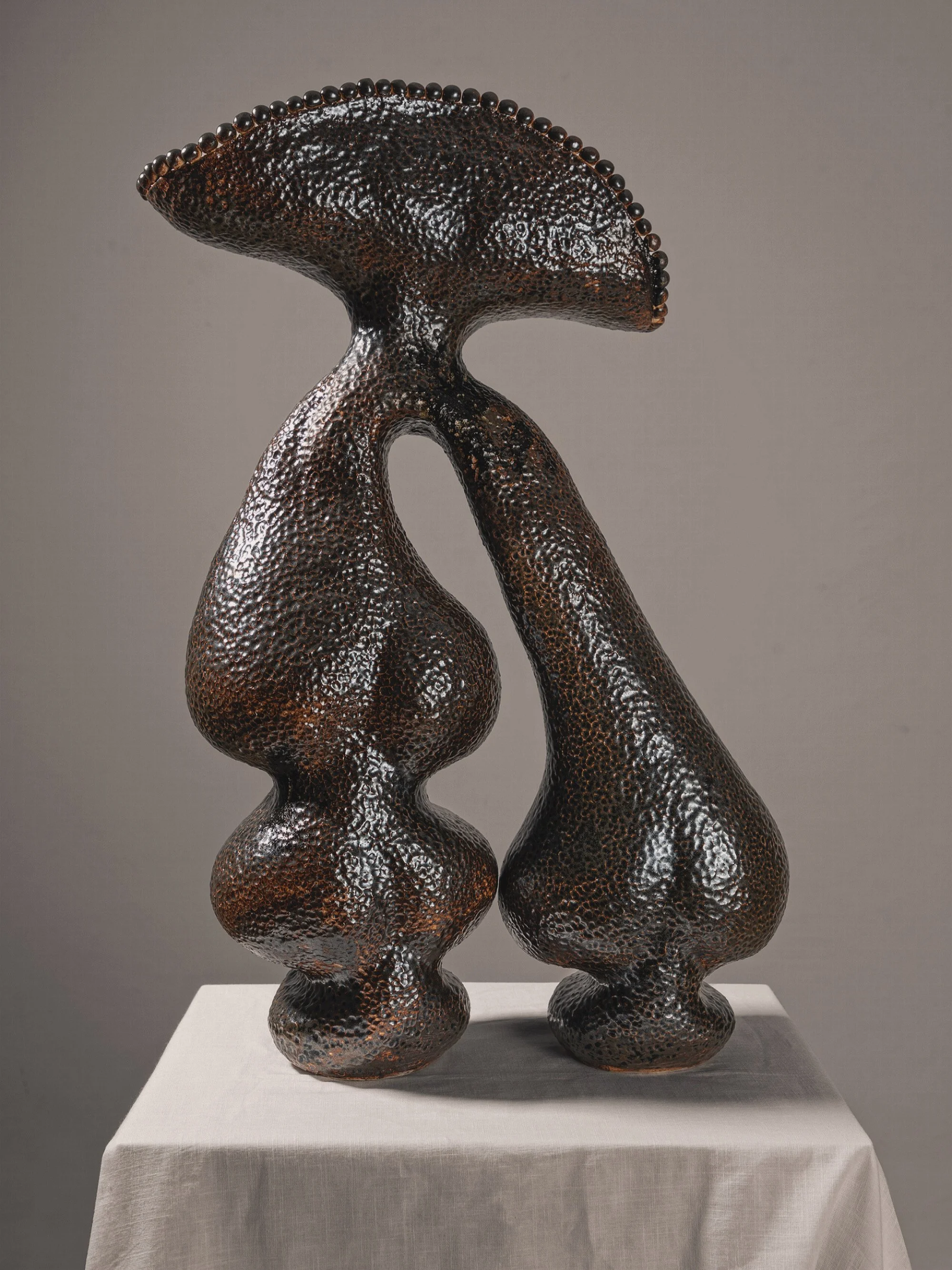 A modern abstract sculpture made of dark, textured metal, displayed on a white pedestal.