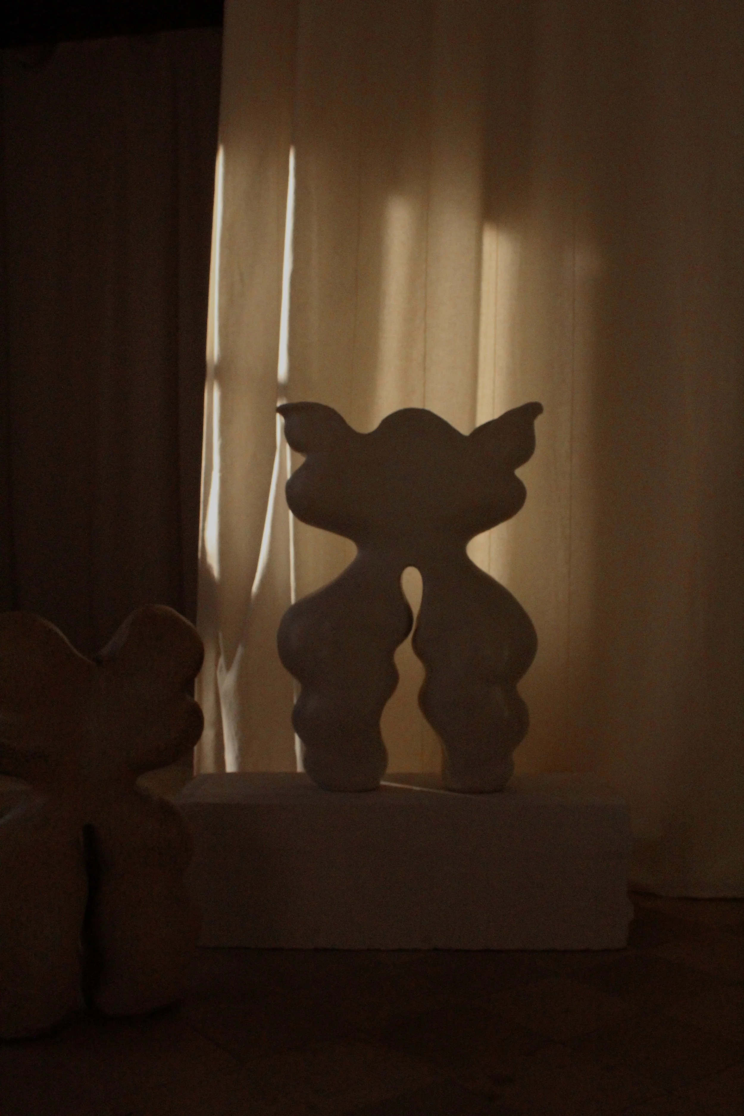 Silhouette of a wooden animal figure, possibly a dog or cat, sitting on a surface in front of a window with curtains, backlit by sunlight.