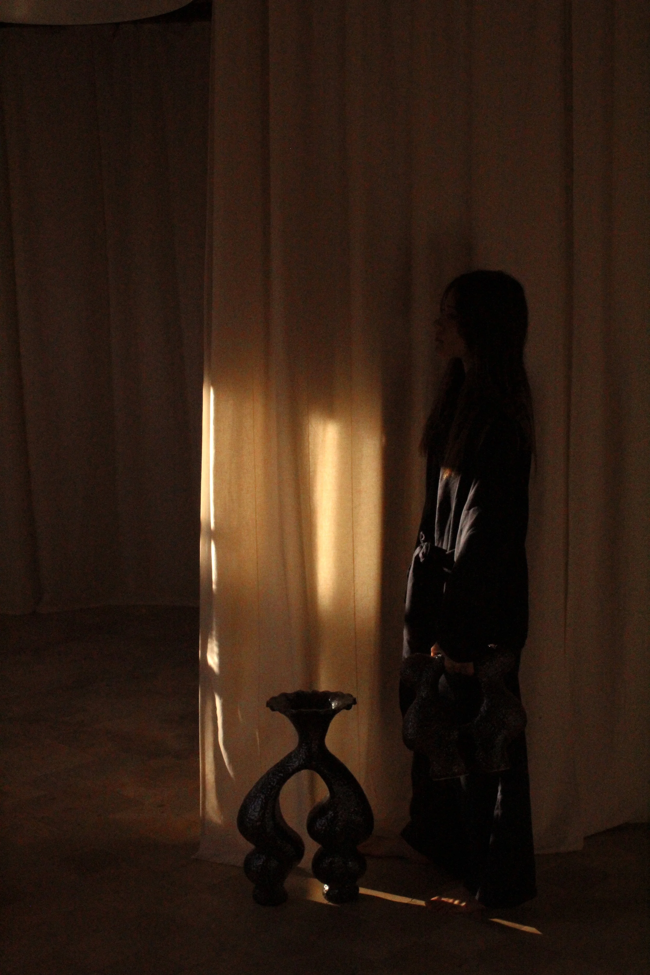 A woman standing against a curtain with sunlight streaming through, holding a bag, with a decorative dark-colored side table on her right.