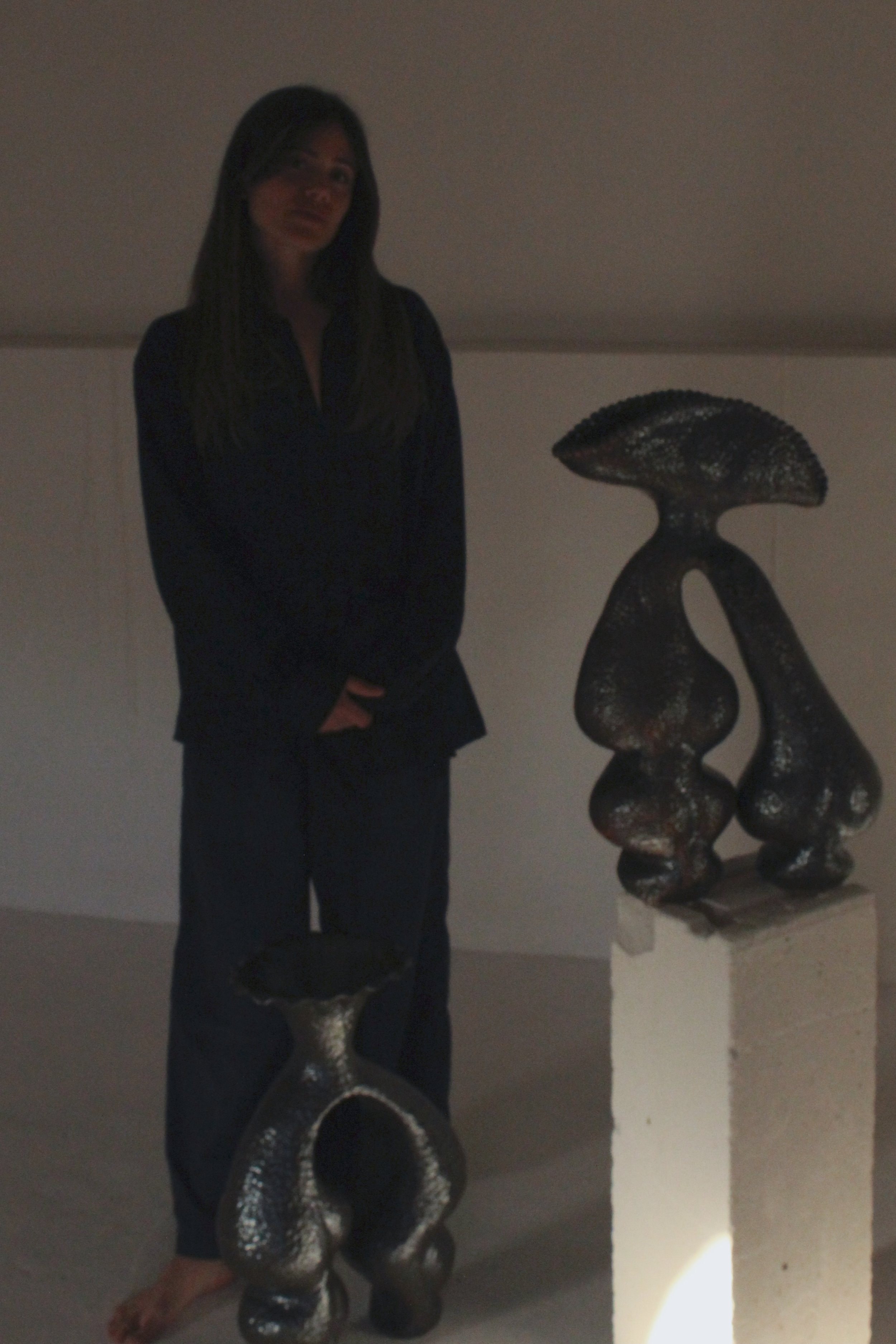 A woman with long dark hair standing next to abstract sculptures on pedestals in a dimly lit space.