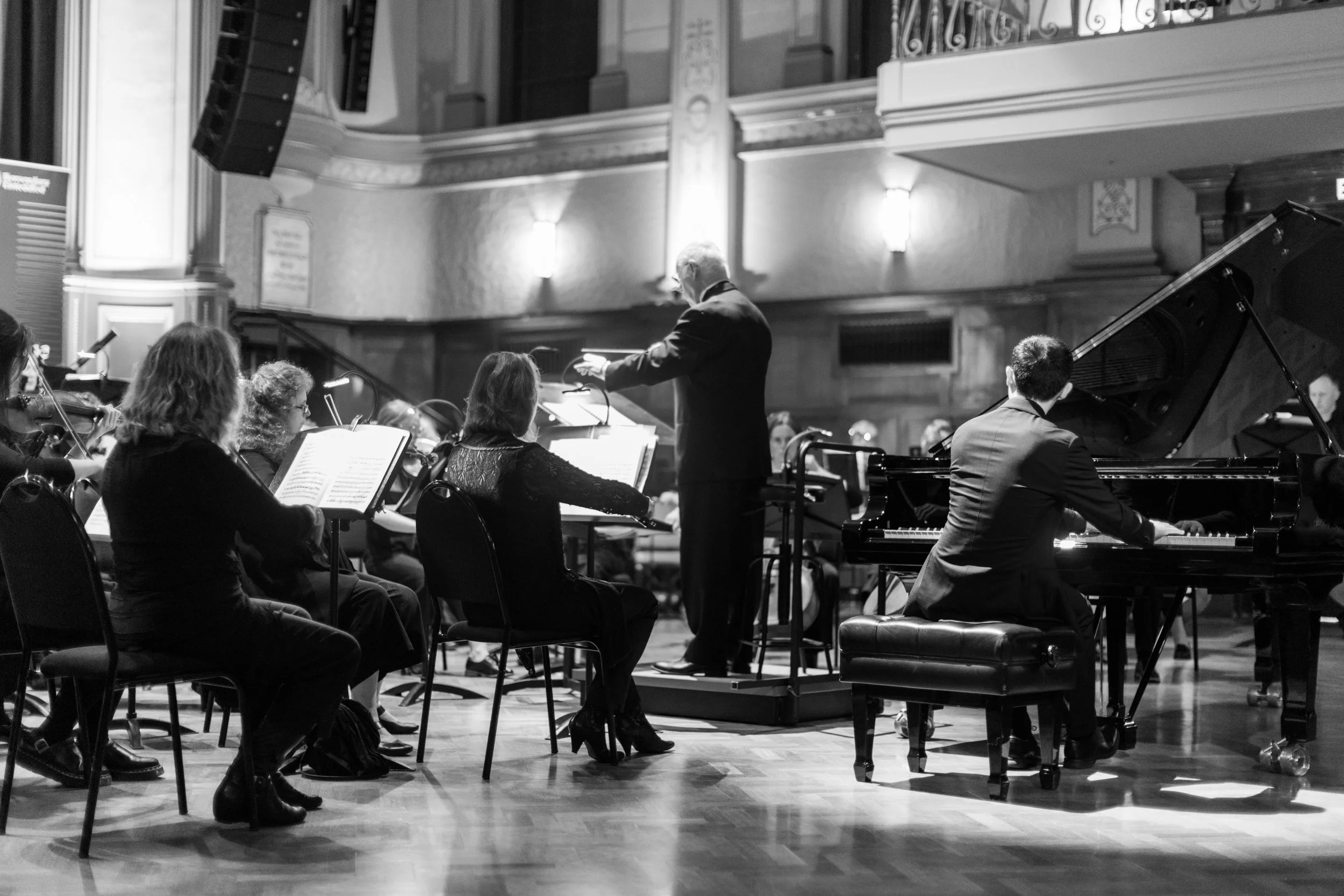 An orchestra performing in a concert hall, with a conductor leading and a pianist playing a grand piano.