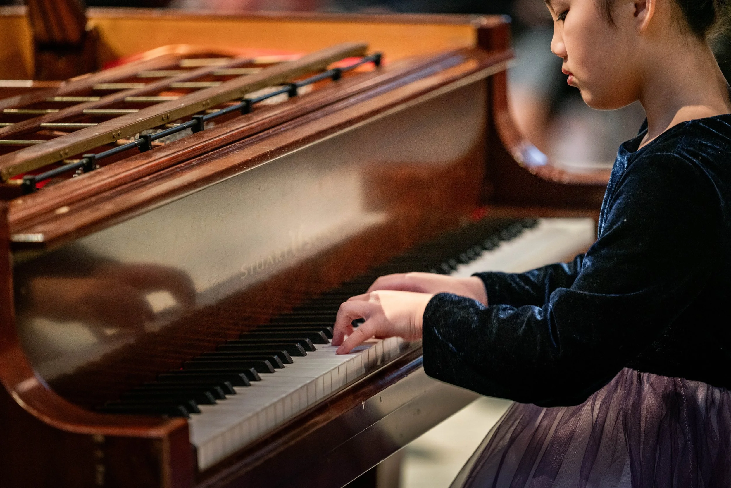 A young girl playing a brown upright piano, wearing a dark velvet long-sleeve top and a pleated skirt.