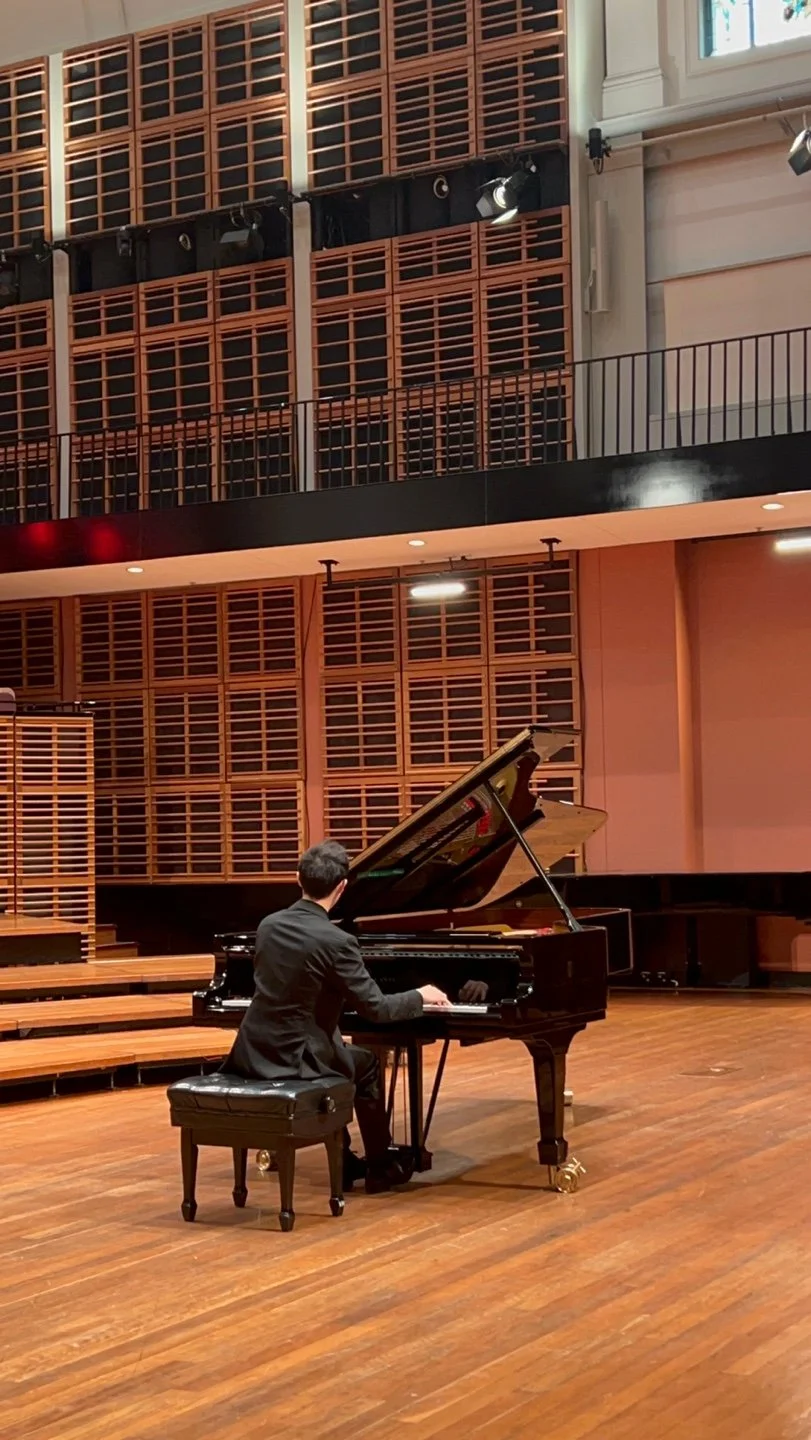 A man in a black suit playing a grand piano on a wooden stage in a hall with wooden paneling and acoustic design elements.