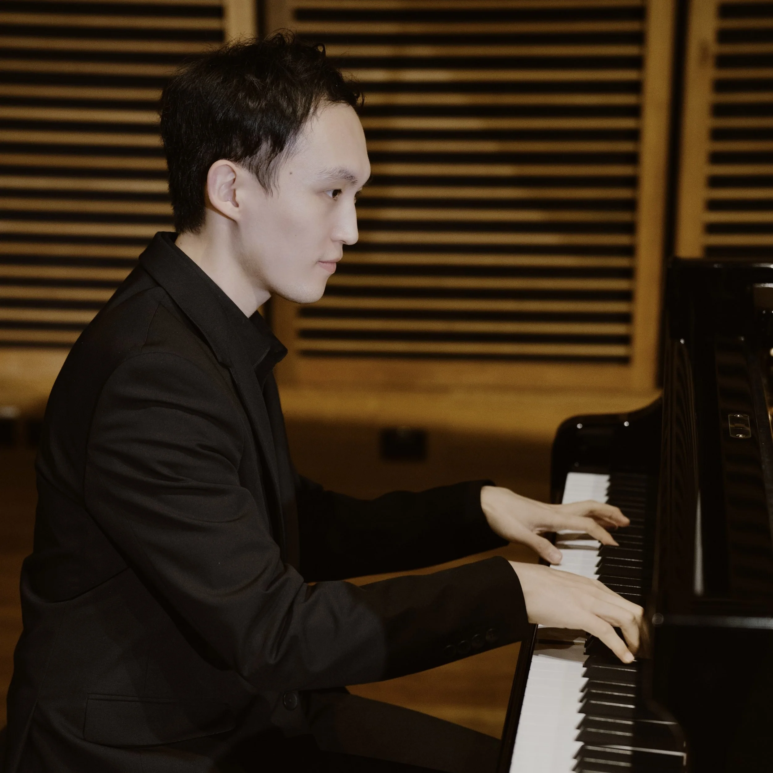 A young man in a black suit playing a grand piano in a room with wooden wall panels.