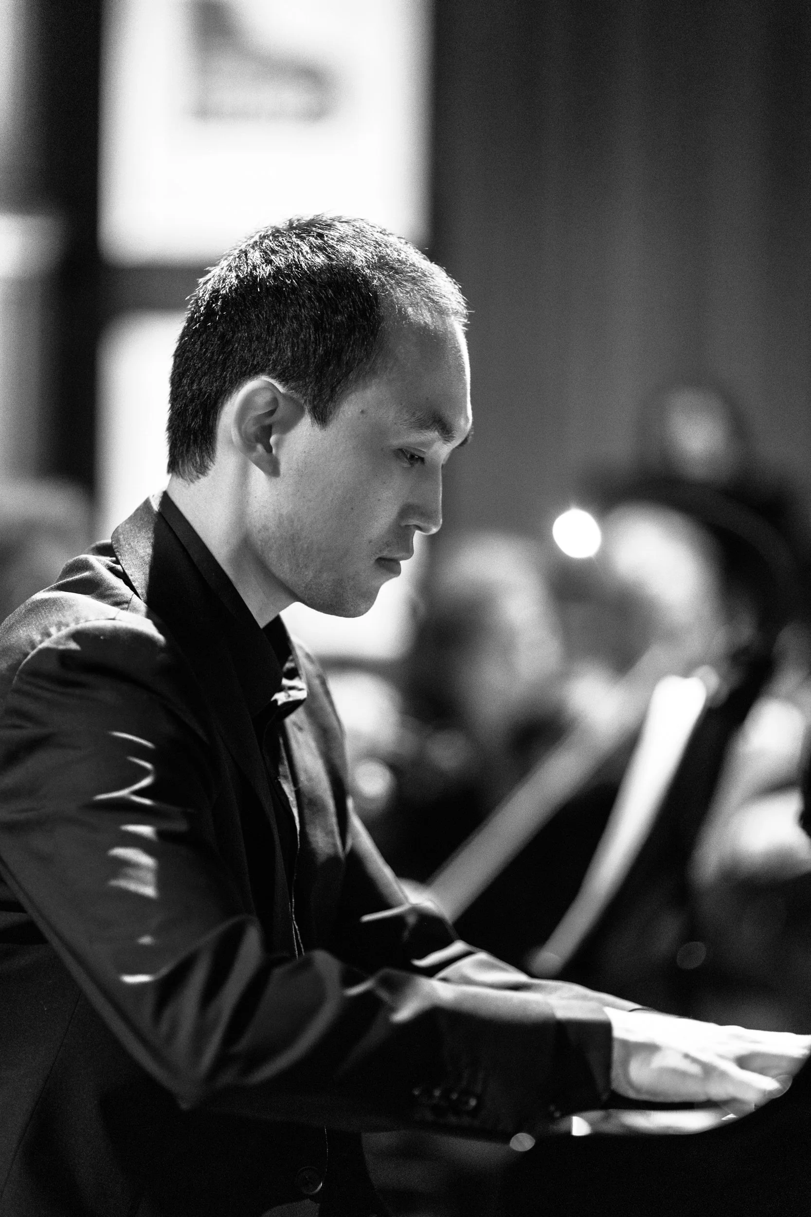 A man with short dark hair is playing the piano, focused on the keys in a black-and-white photo.