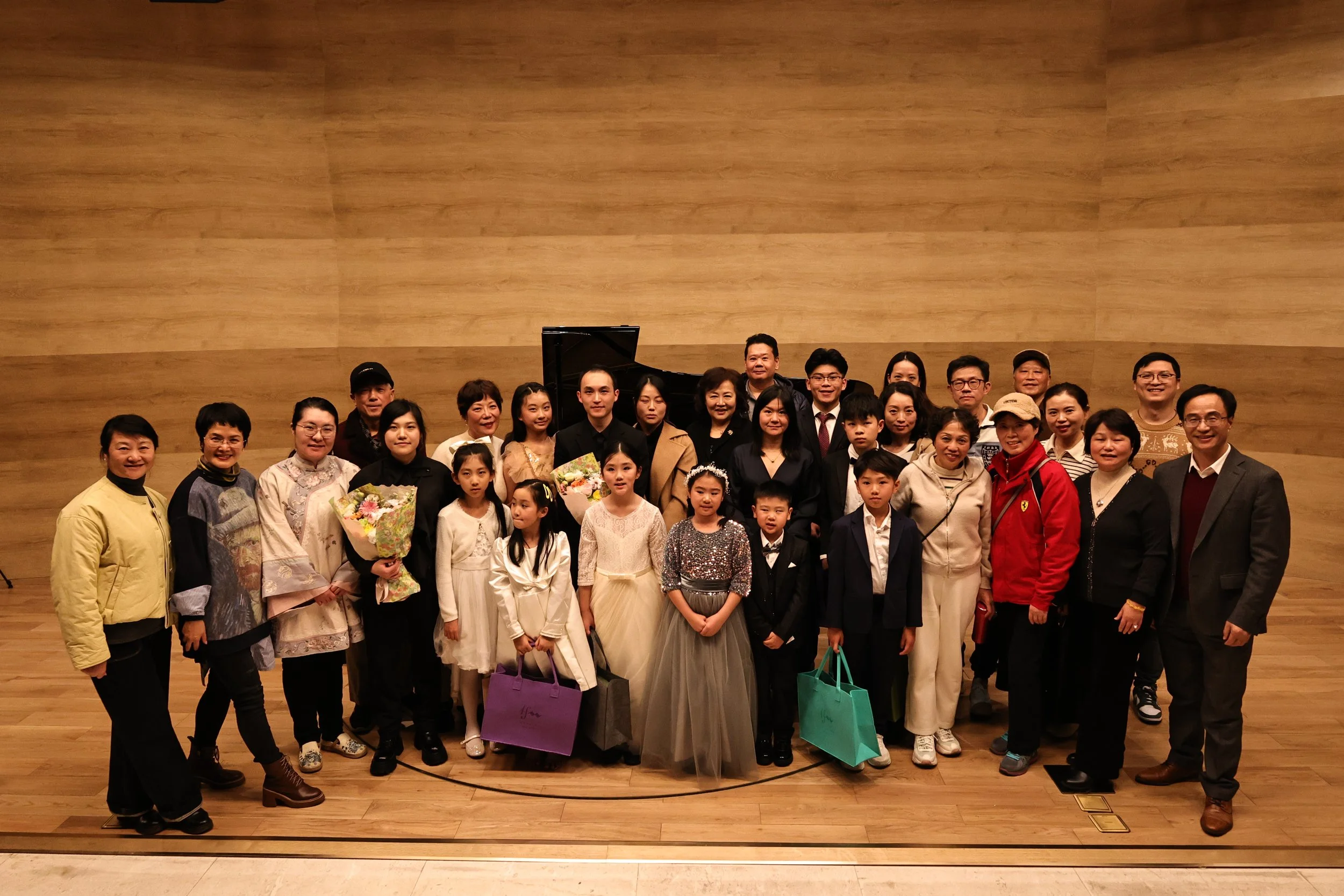 A large group of people, including children and adults, posing together on a stage with a piano in the background. Some people are holding flowers and gift bags.