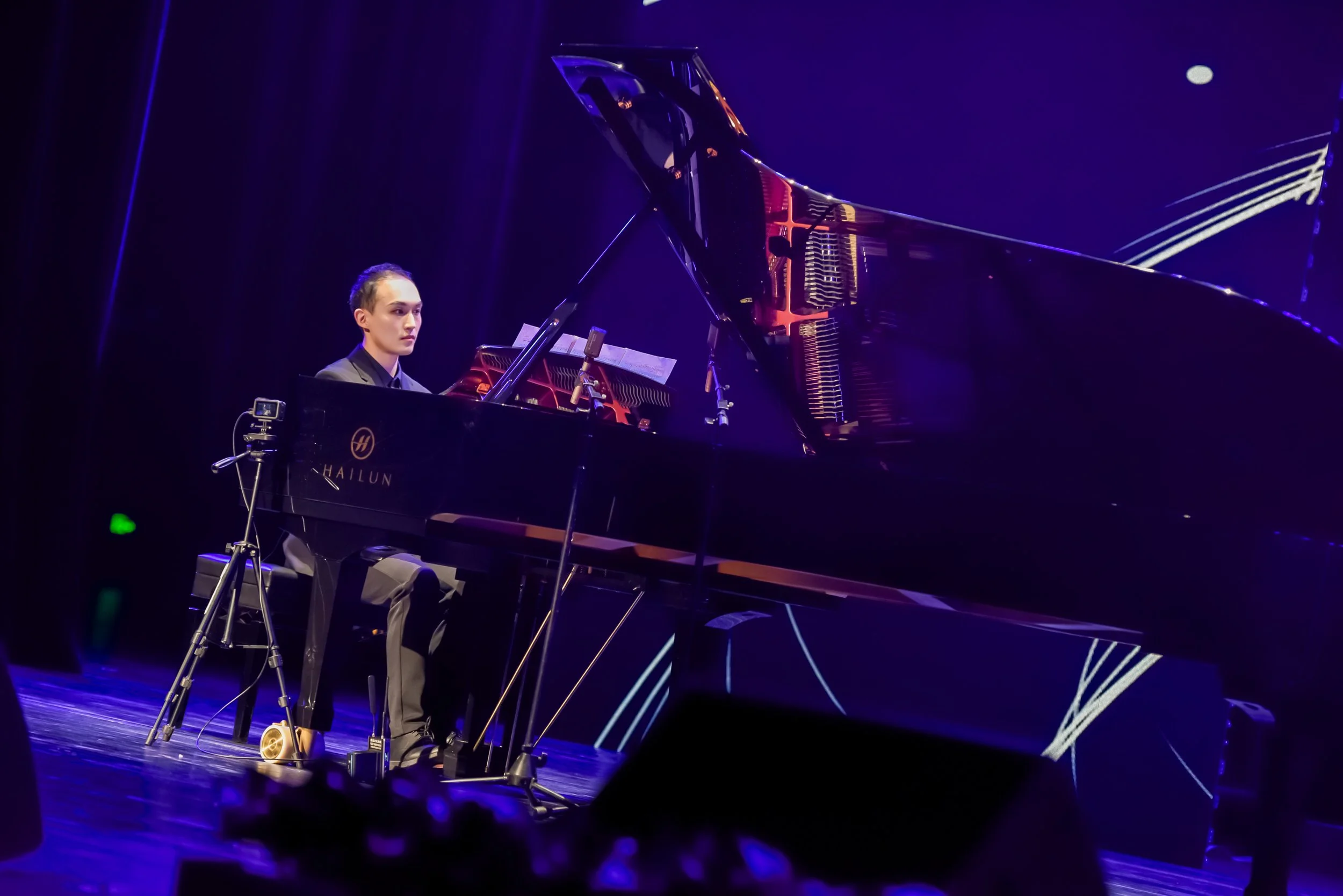 A person playing a grand piano on stage during a performance, with a dark background and stage lighting.
