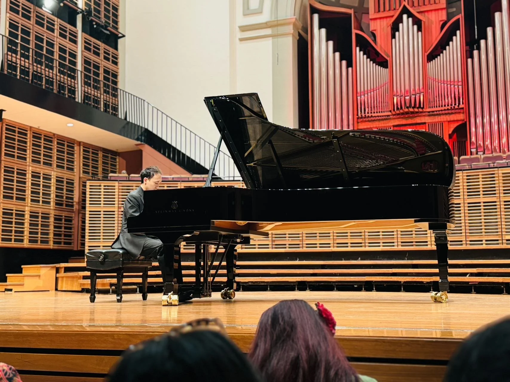 A person playing a grand piano on stage in a concert hall surrounded by wooden acoustical panels, with an audience visible in the foreground.