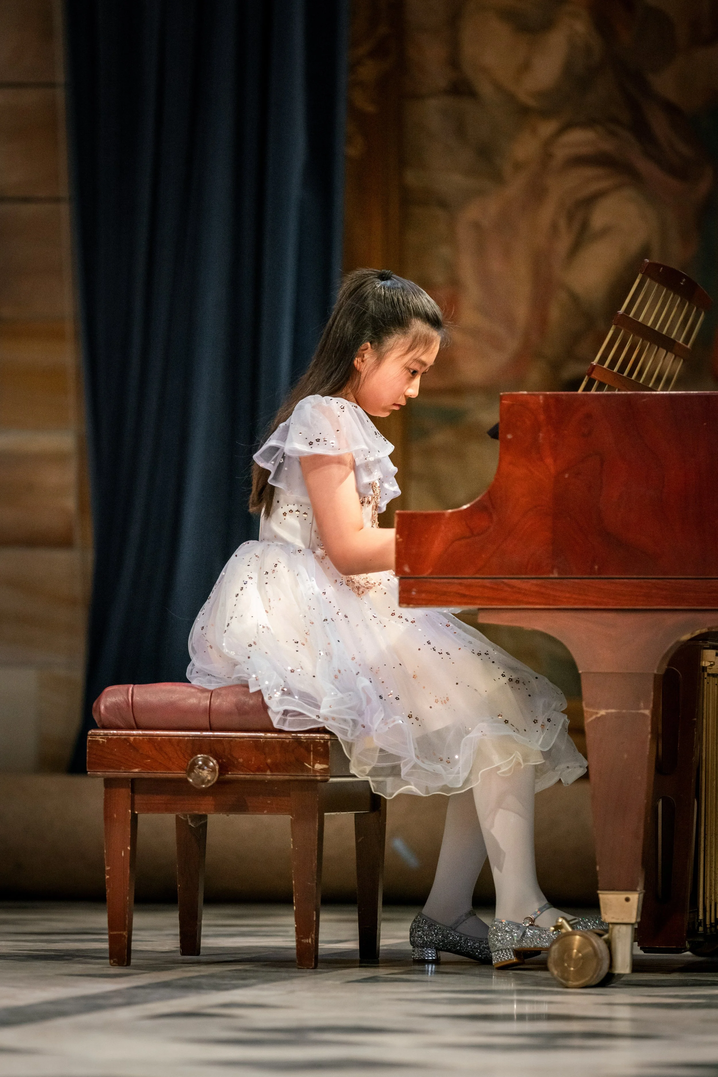 A young girl in a white dress with glitter details seated at a grand piano, focusing on playing during a performance or recital.