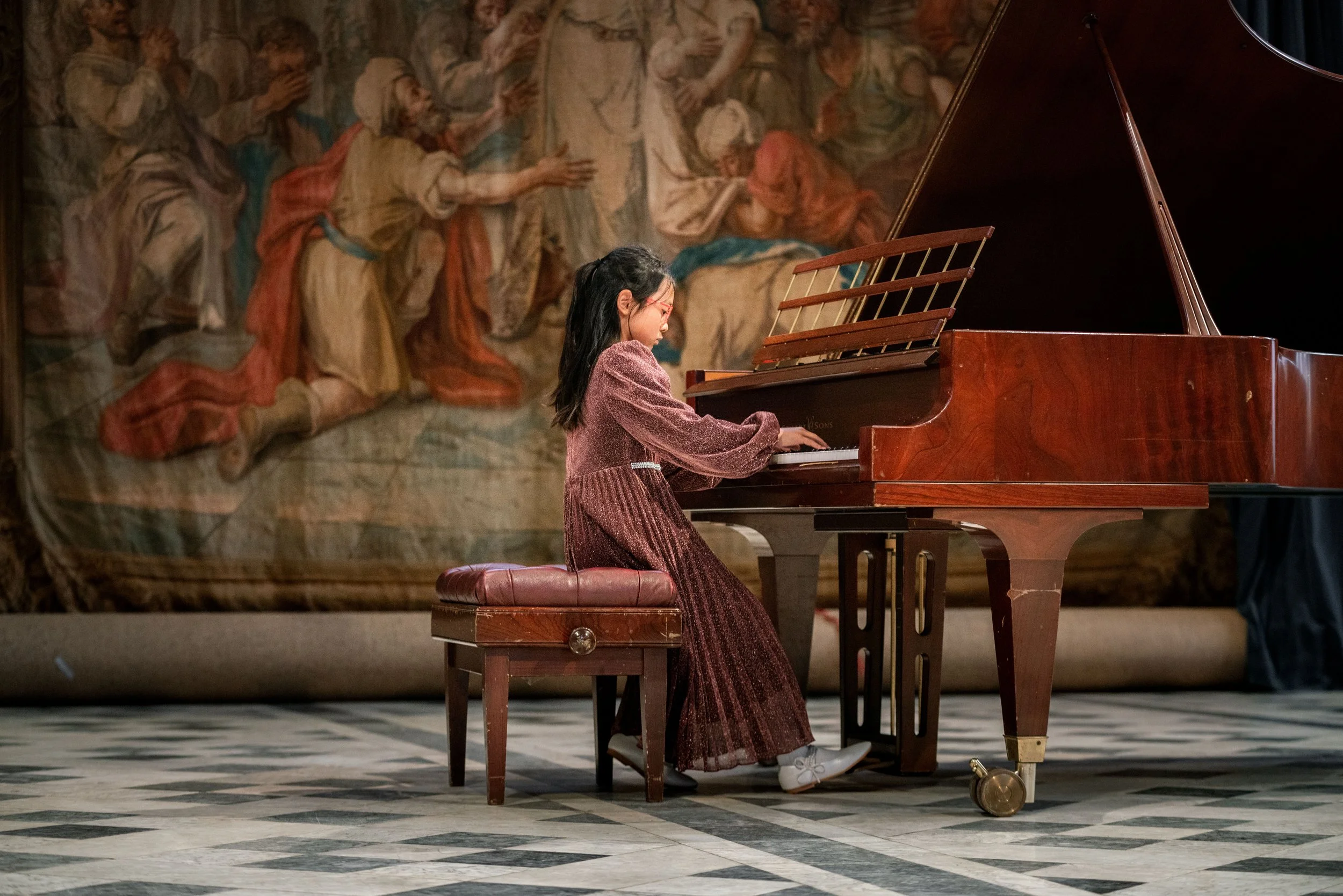 A young girl playing the piano on a stage with a historical painting in the background.