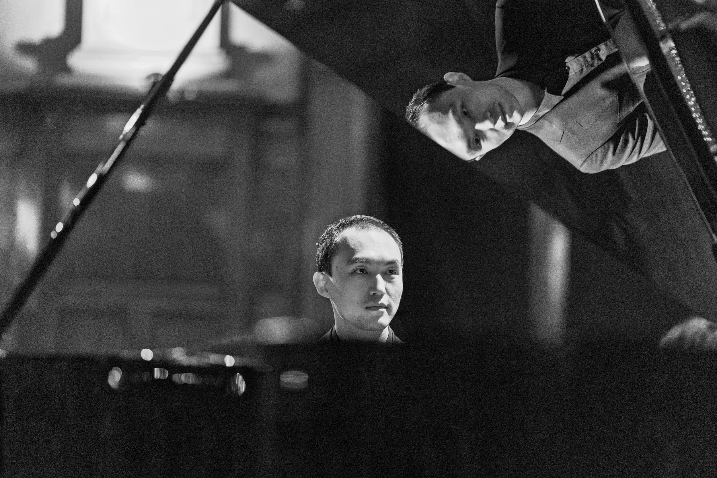 Two men playing piano in a room, one of them is looking at the camera, the other is focusing on the piano keys.