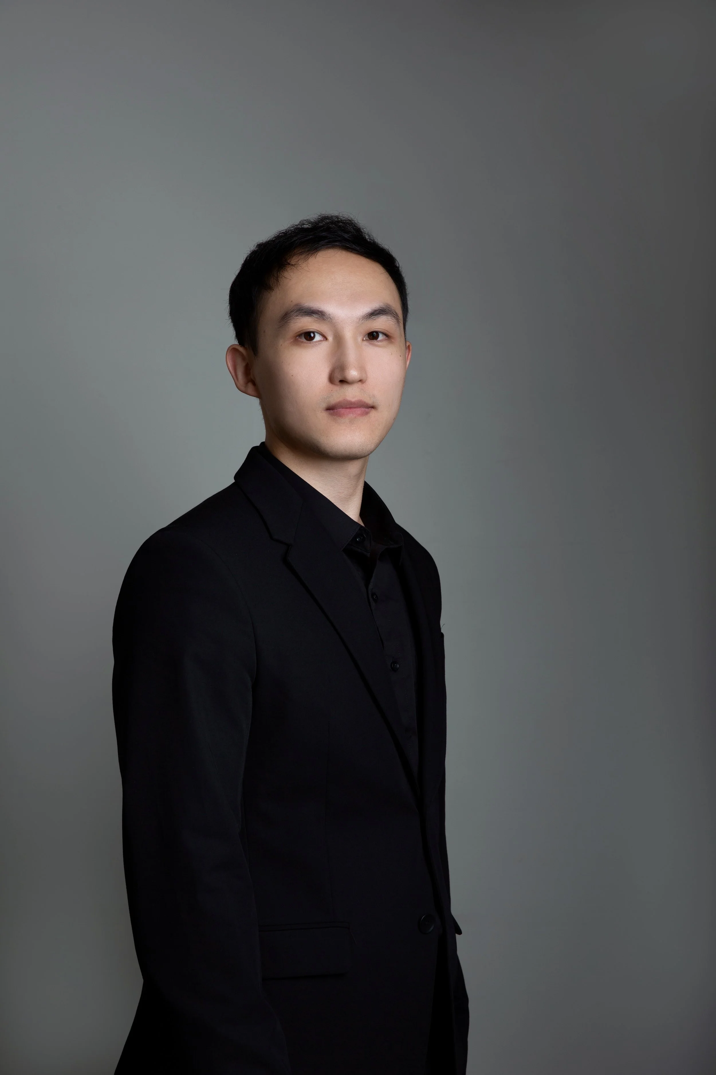 A young man with black hair and light skin wearing a black suit and shirt standing against a gray background.