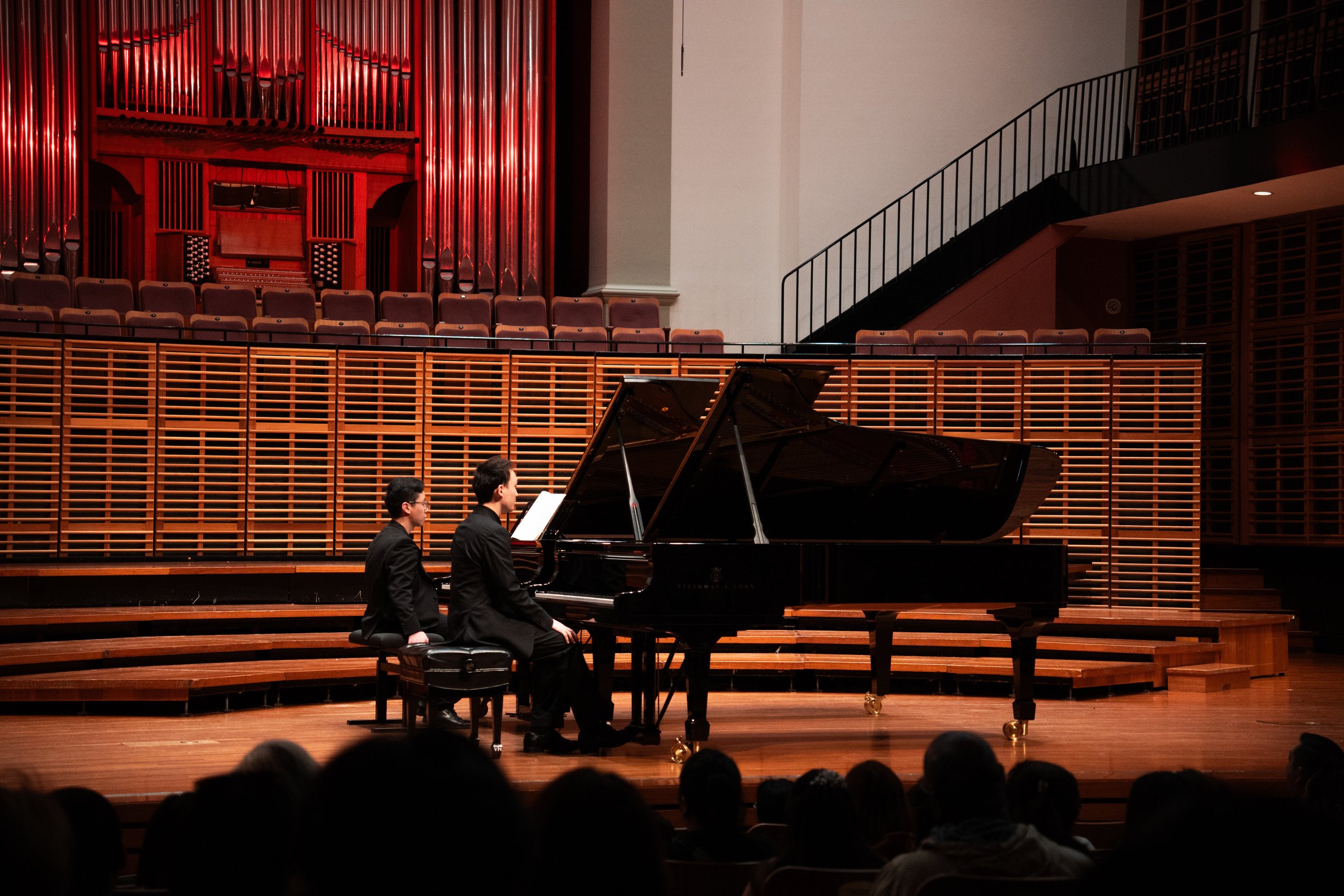 Two musicians in formal black attire playing a grand piano on a stage in a concert hall, with audience members visible in the foreground.